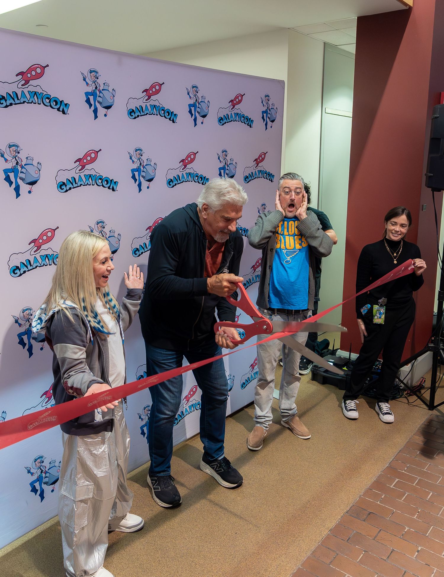 Celebrities cutting a red ribbon at a galaxycon event with cartoon character wall decorations.