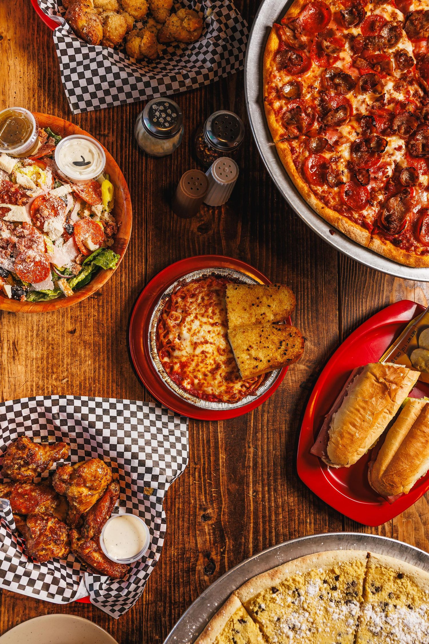 Overhead view of a restaurant spread featuring pepperoni pizza, wings, lasagna, breadsticks, salad, and fried appetizers on wood table.