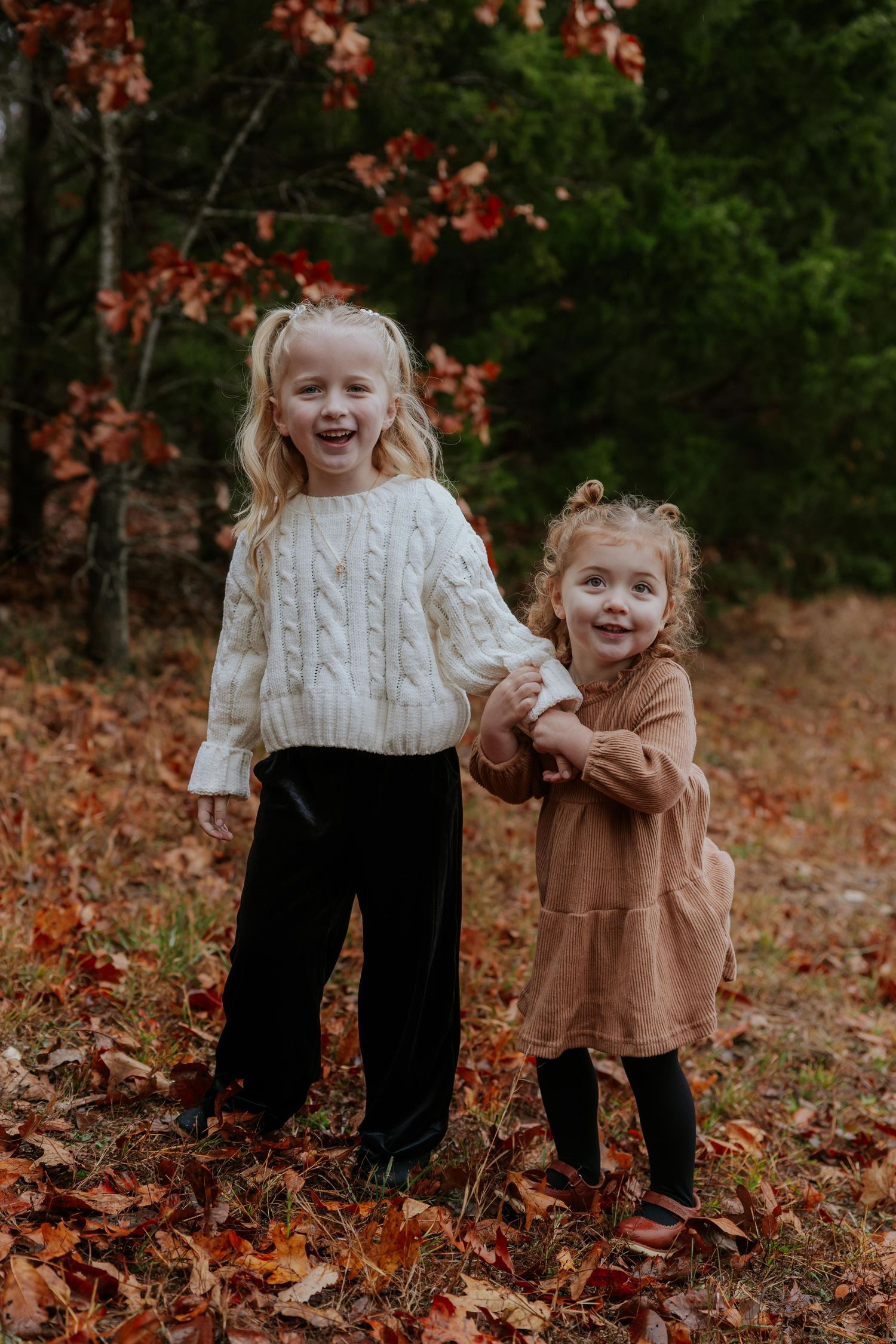 Two children in sweaters holding hands and laughing among fallen autumn leaves in Missouri.