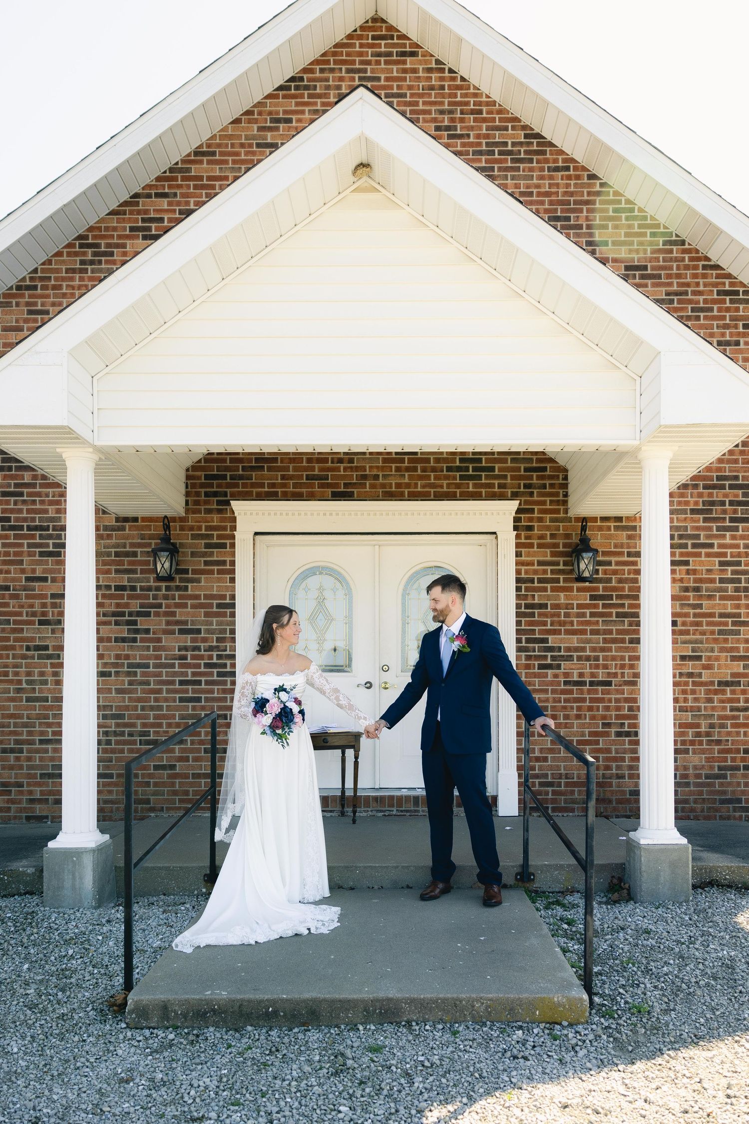 Bride and groom holding hands on church steps, she in white gown with bouquet, he in navy suit, brick chapel backdrop.
