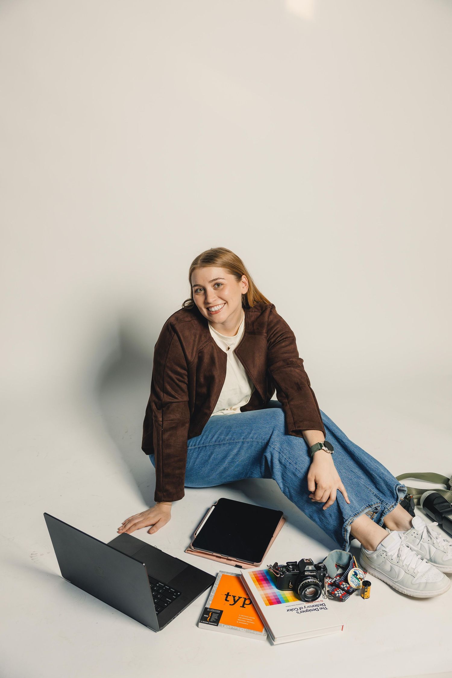 Smiling young woman sitting on white floor with laptop, tablet, camera, and colorful books around her.