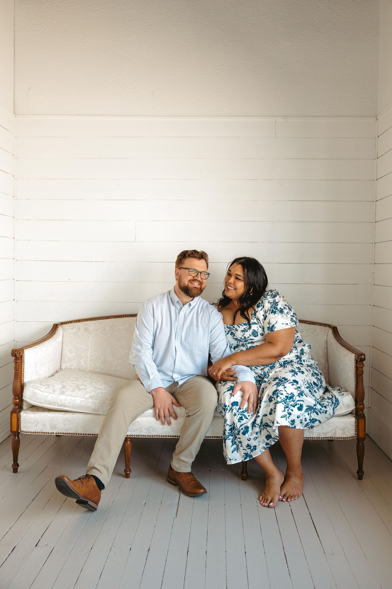 Happy interracial couple sitting closely together on a vintage white sofa in a bright white room.