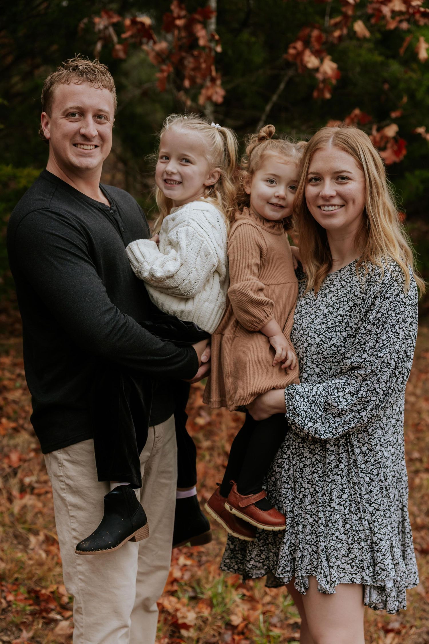 A family of four poses together outdoors in autumn with fallen leaves and colorful foliage in the background.
