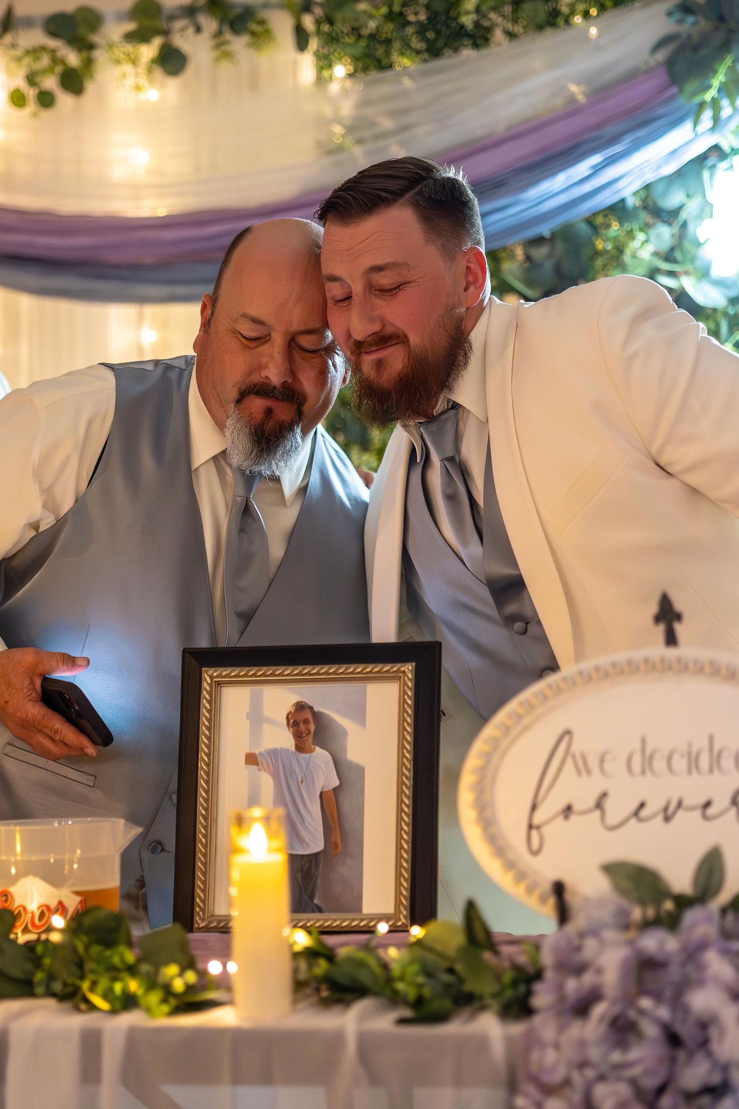 Father and son embrace at a wedding reception table decorated with candles, greenery and purple accents.