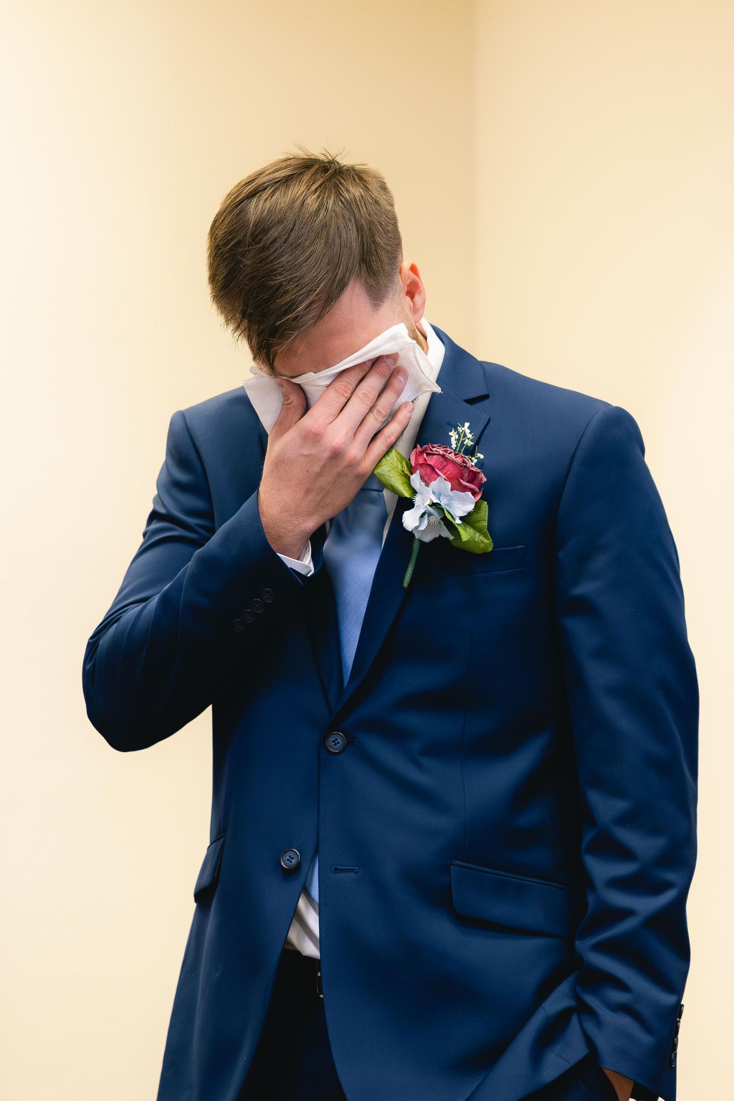 Emotional groom in navy suit wiping tears with handkerchief, wearing colorful boutonniere on wedding day.