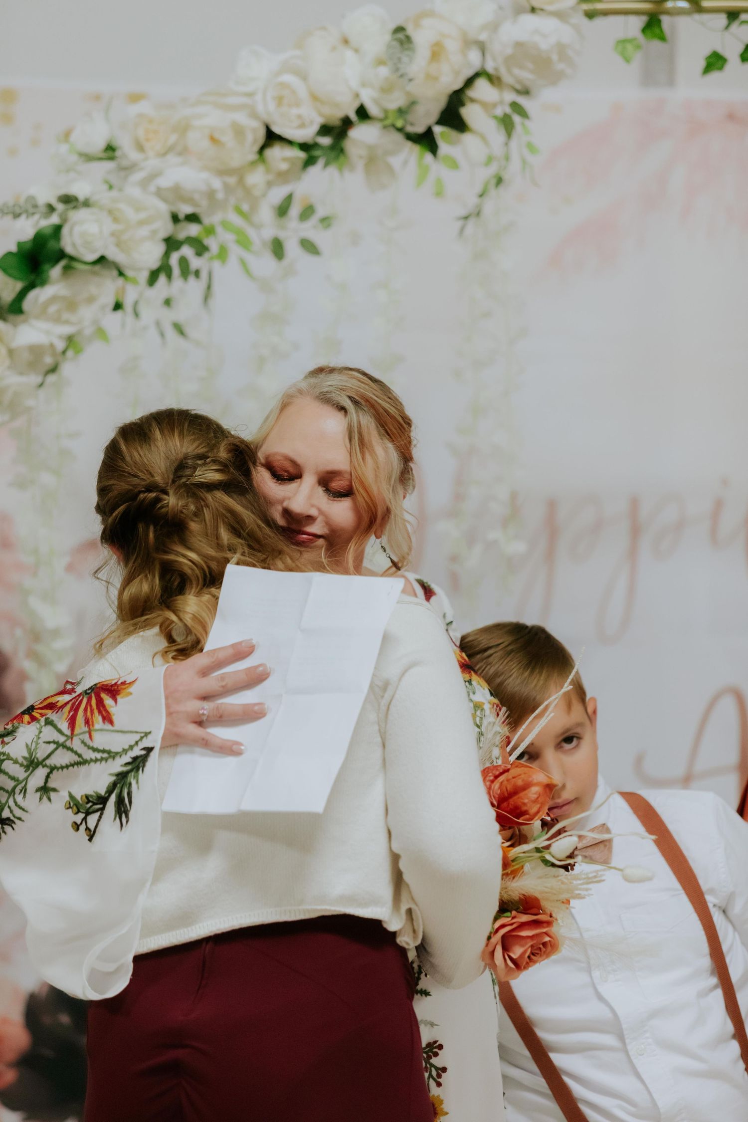 Emotional wedding ceremony moment captured under white floral arch with burgundy accents.