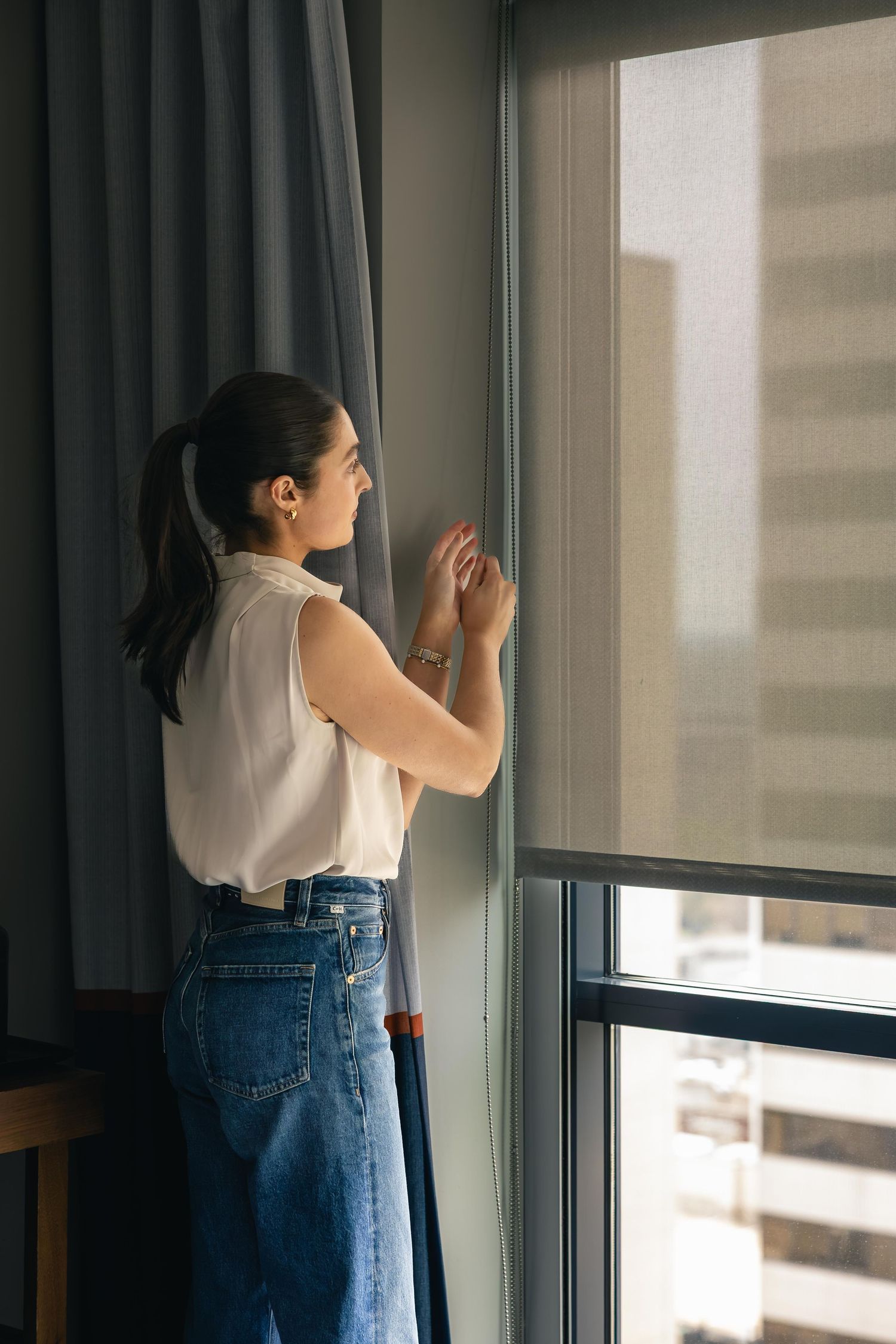 Woman in white blouse and jeans adjusting window blinds while gazing outside in a modern room.