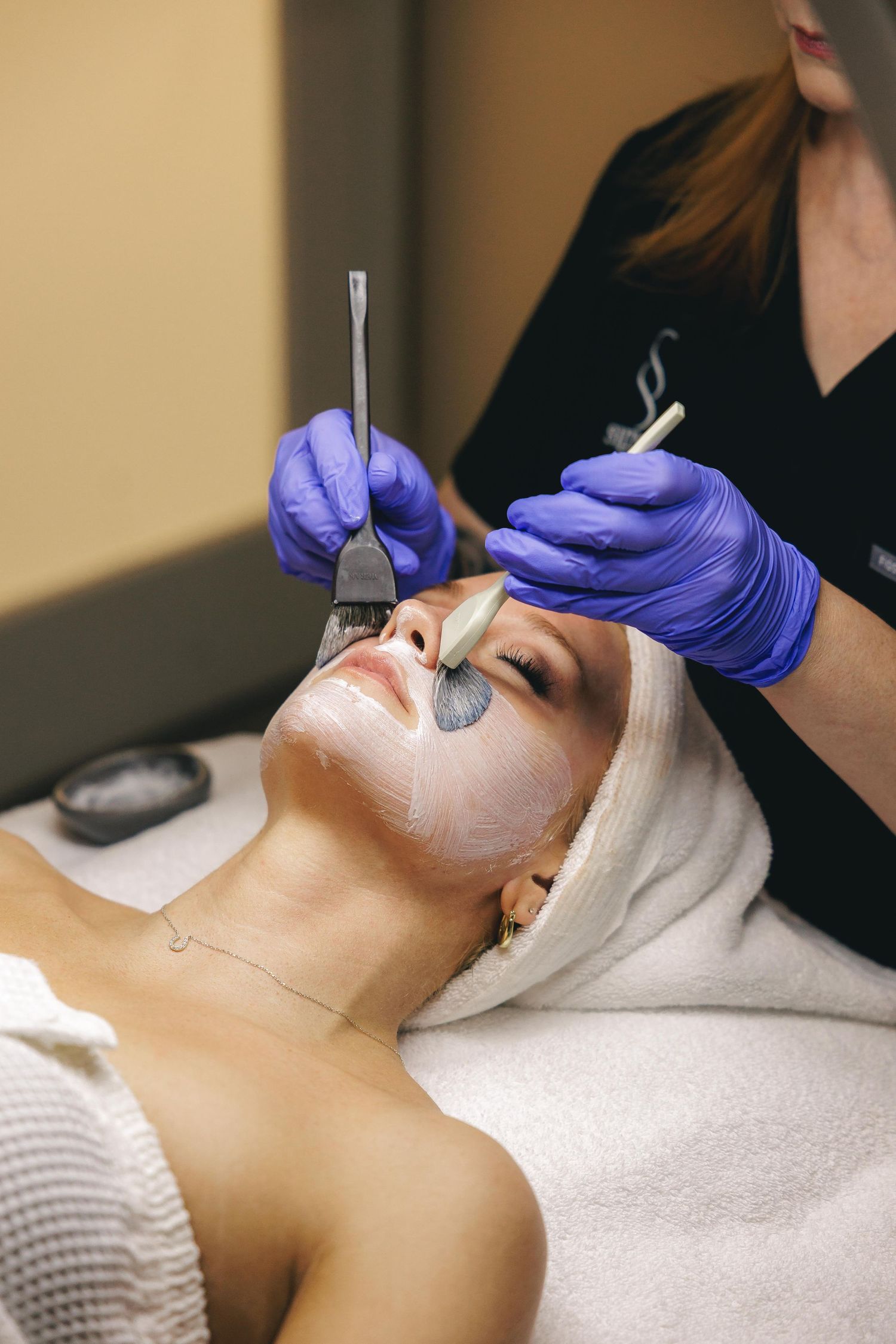 Esthetician applying dual-tone facial mask with brushes on relaxed woman lying on spa treatment table.