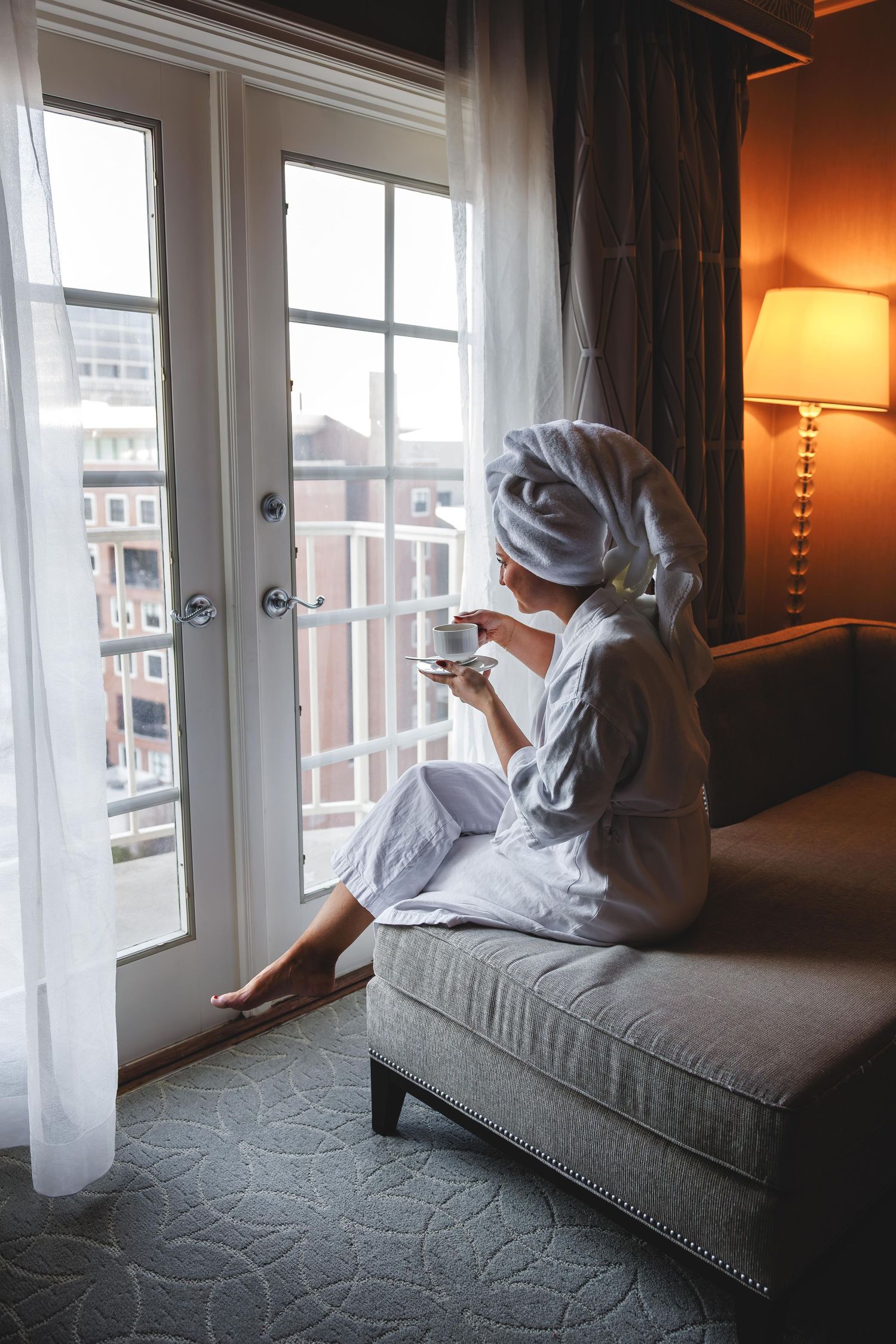 Woman in white bathrobe and hair towel sitting on sofa, enjoying coffee while gazing out hotel room window.