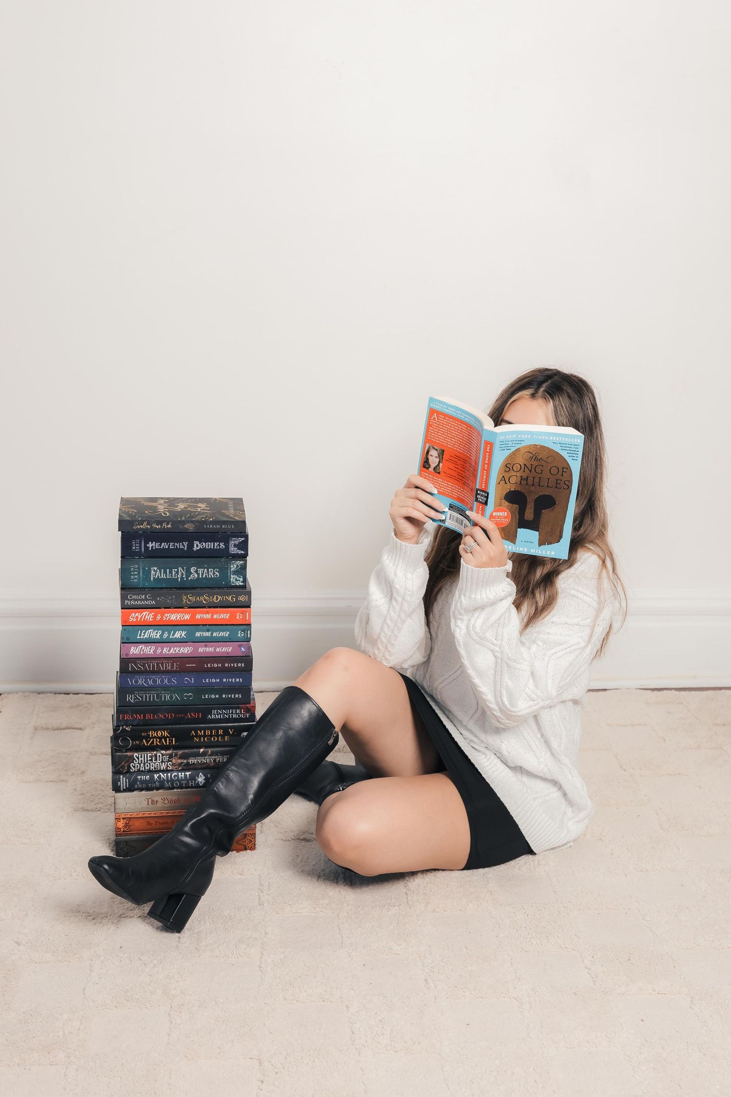 Young woman in white sweater and black boots sits on floor reading a book beside a tall stack of novels.
