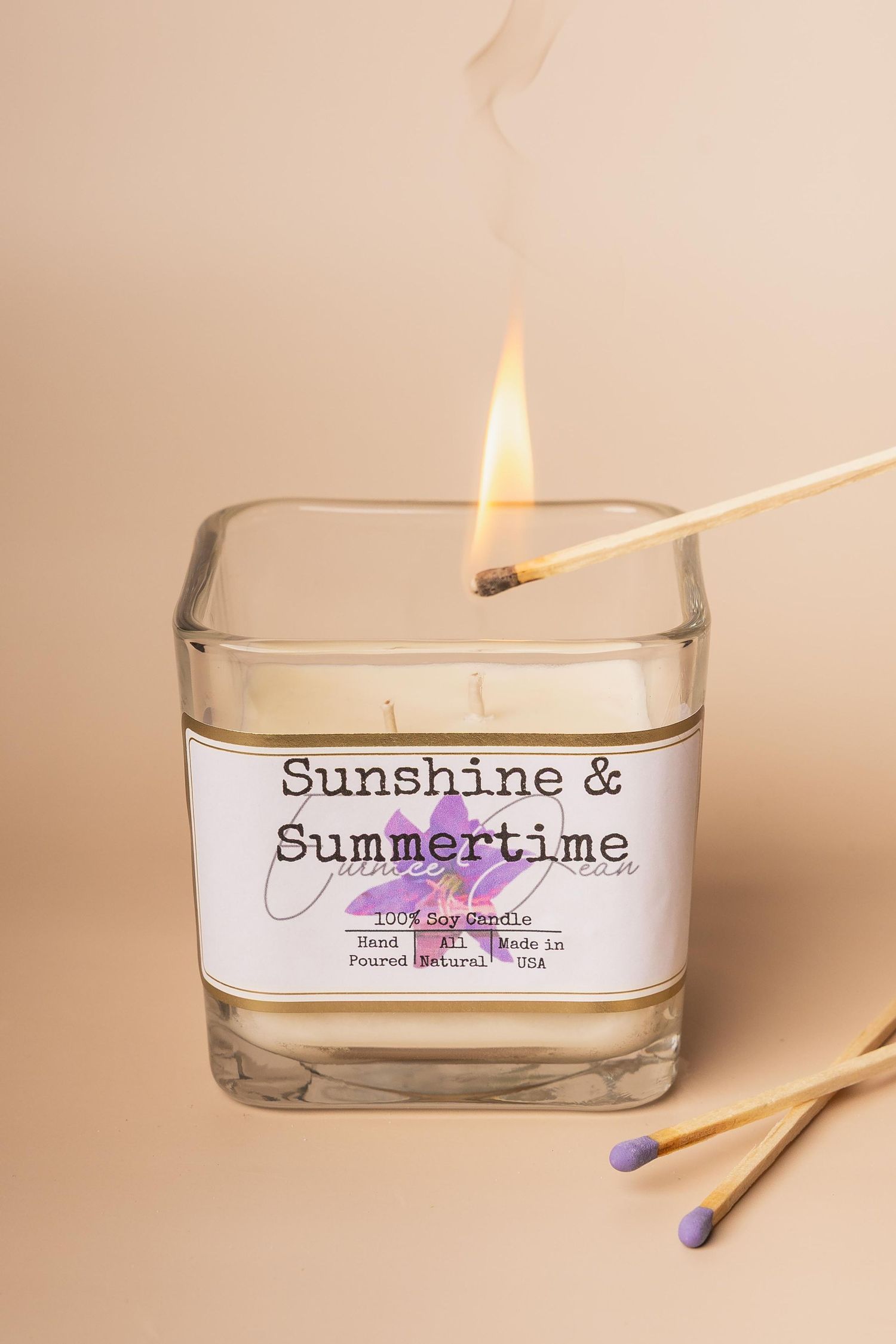 A lit Sunshine & Summertime soy candle in a glass jar being ignited by a match on a beige background.