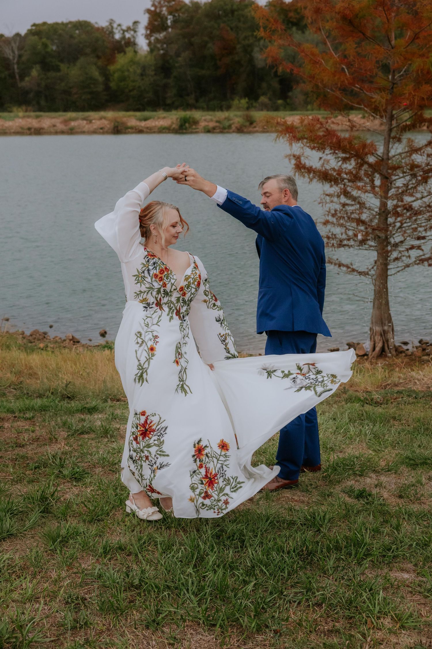 Couple dances playfully on grassy shore with lake and autumn trees in background.
