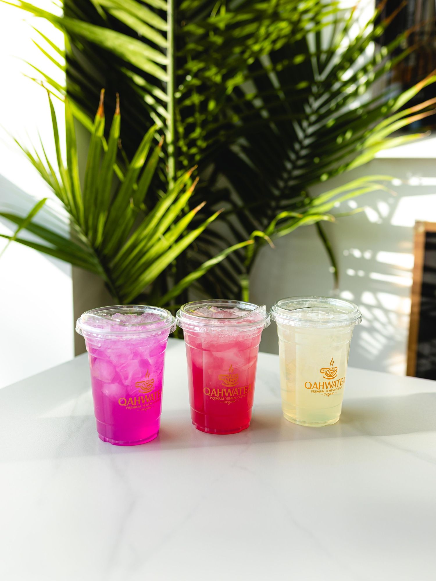 Three colorful tropical drinks in clear plastic cups with ice, featuring vibrant pink and yellow beverages against palm leaves.