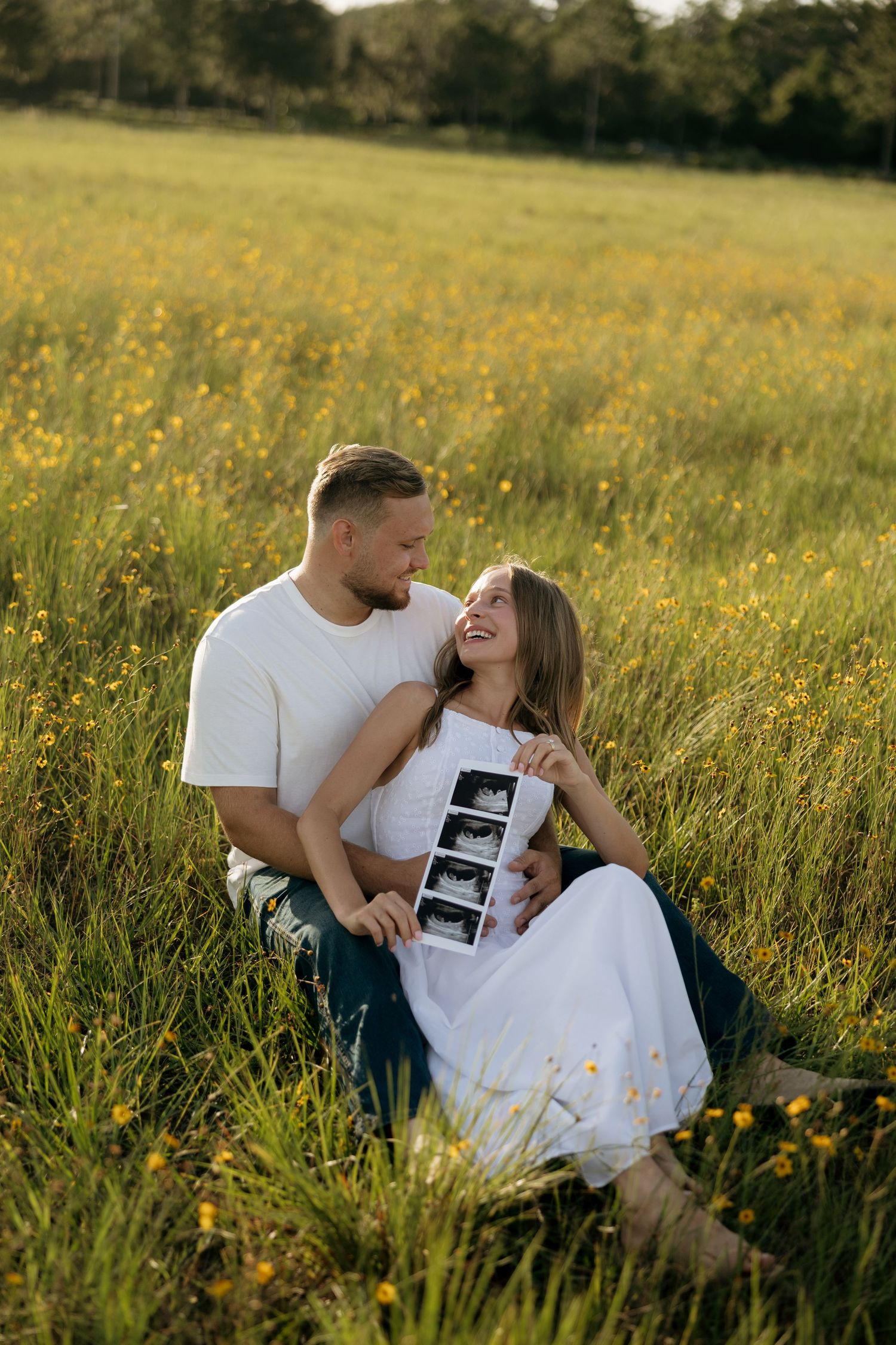 Happy couple sitting in a sunny wildflower field, holding ultrasound photos, announcing their pregnancy.