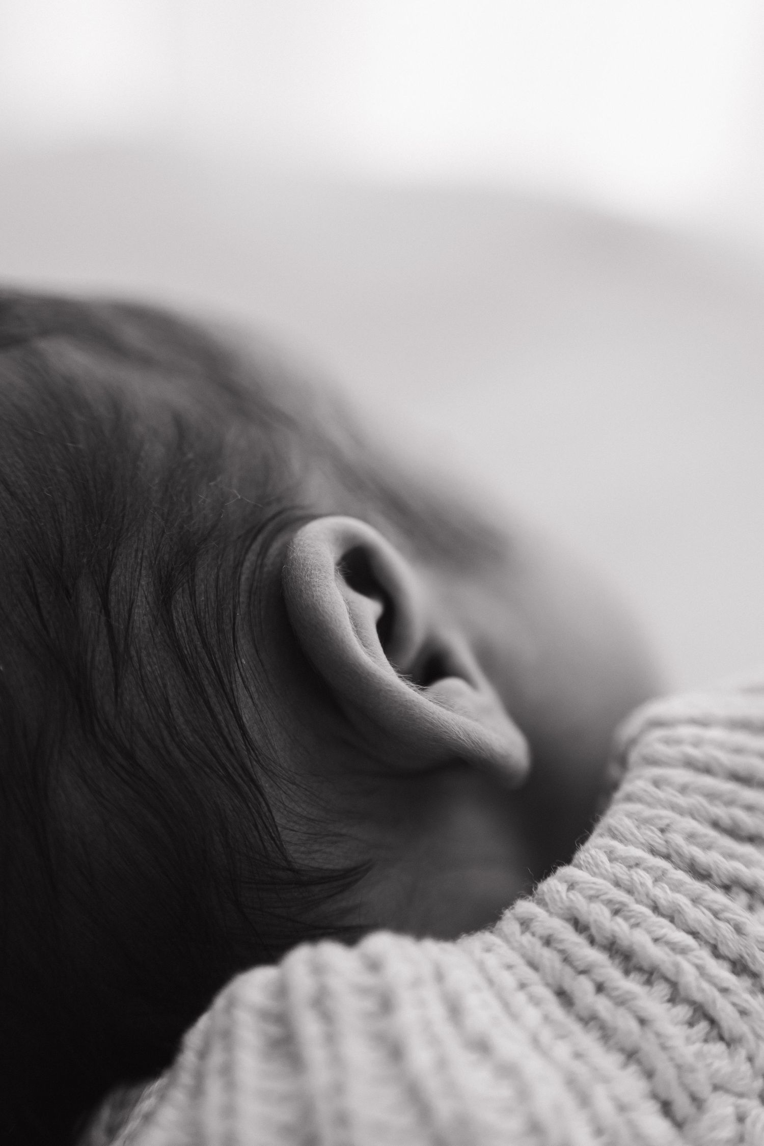 Close-up black and white photo of a newborn baby's ear with dark hair and knit blanket.
