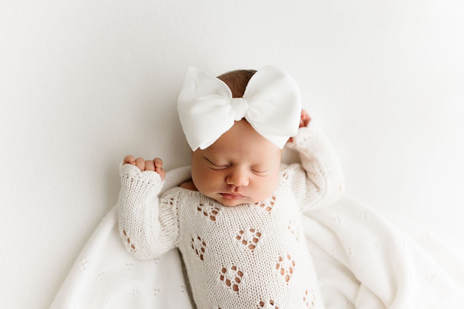 Sleeping newborn baby in white knit outfit and bow headband on white background.