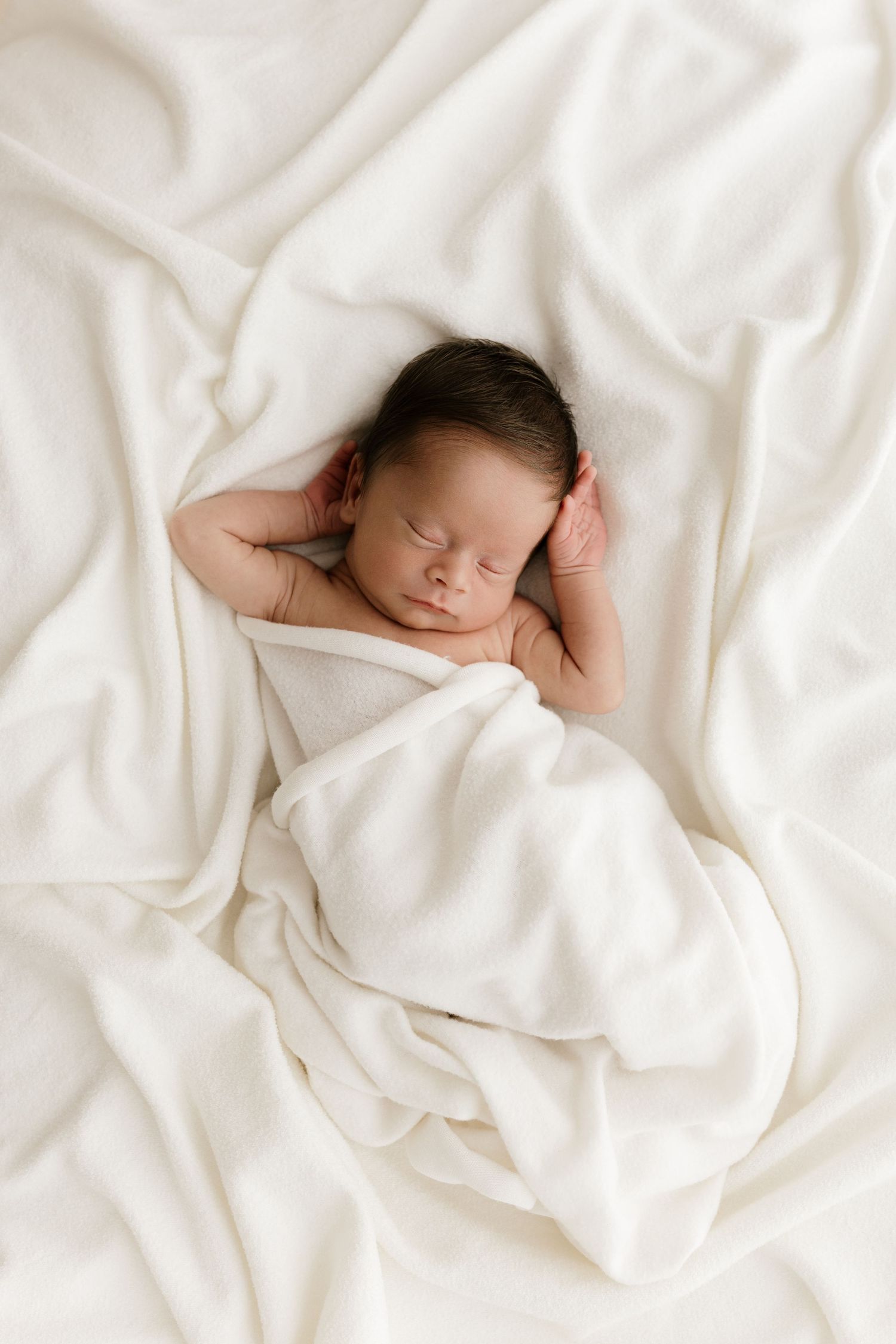 Peaceful newborn baby sleeping wrapped in a soft white blanket on a white bedsheet.