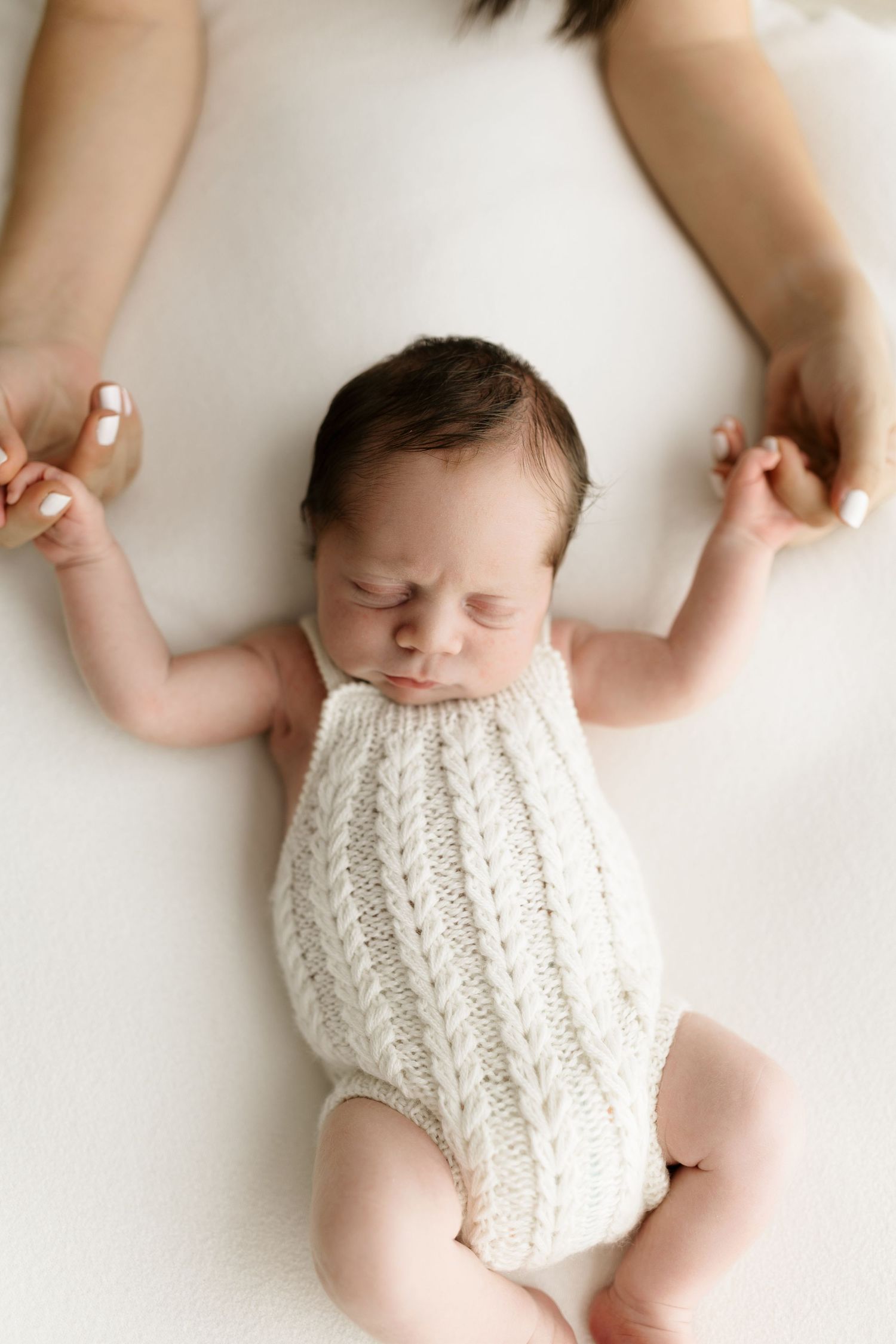 Newborn baby in cream knit romper held by mother's hands on white background.
