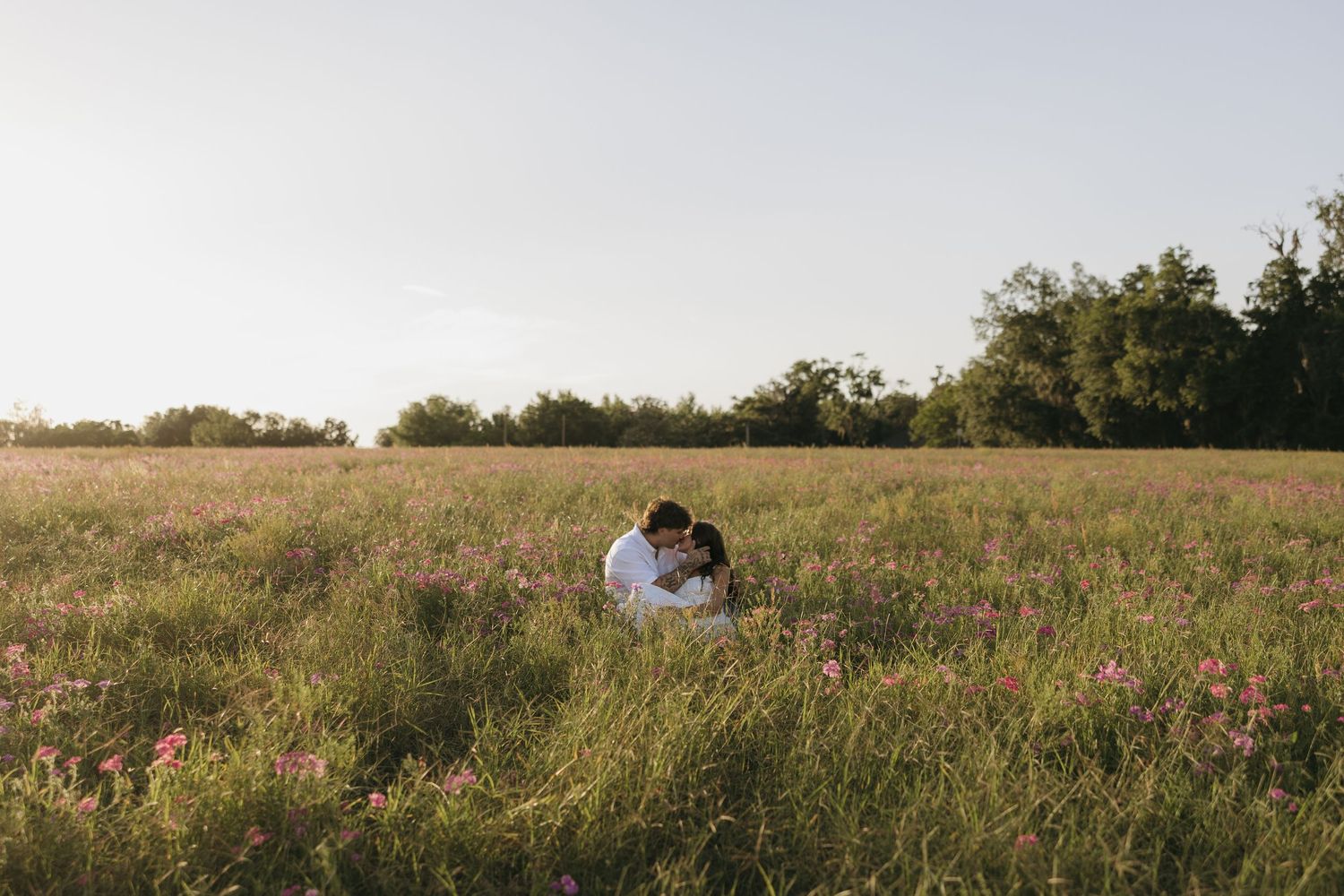 Couple embracing in a lush wildflower field at golden hour with trees and soft sky in the background.