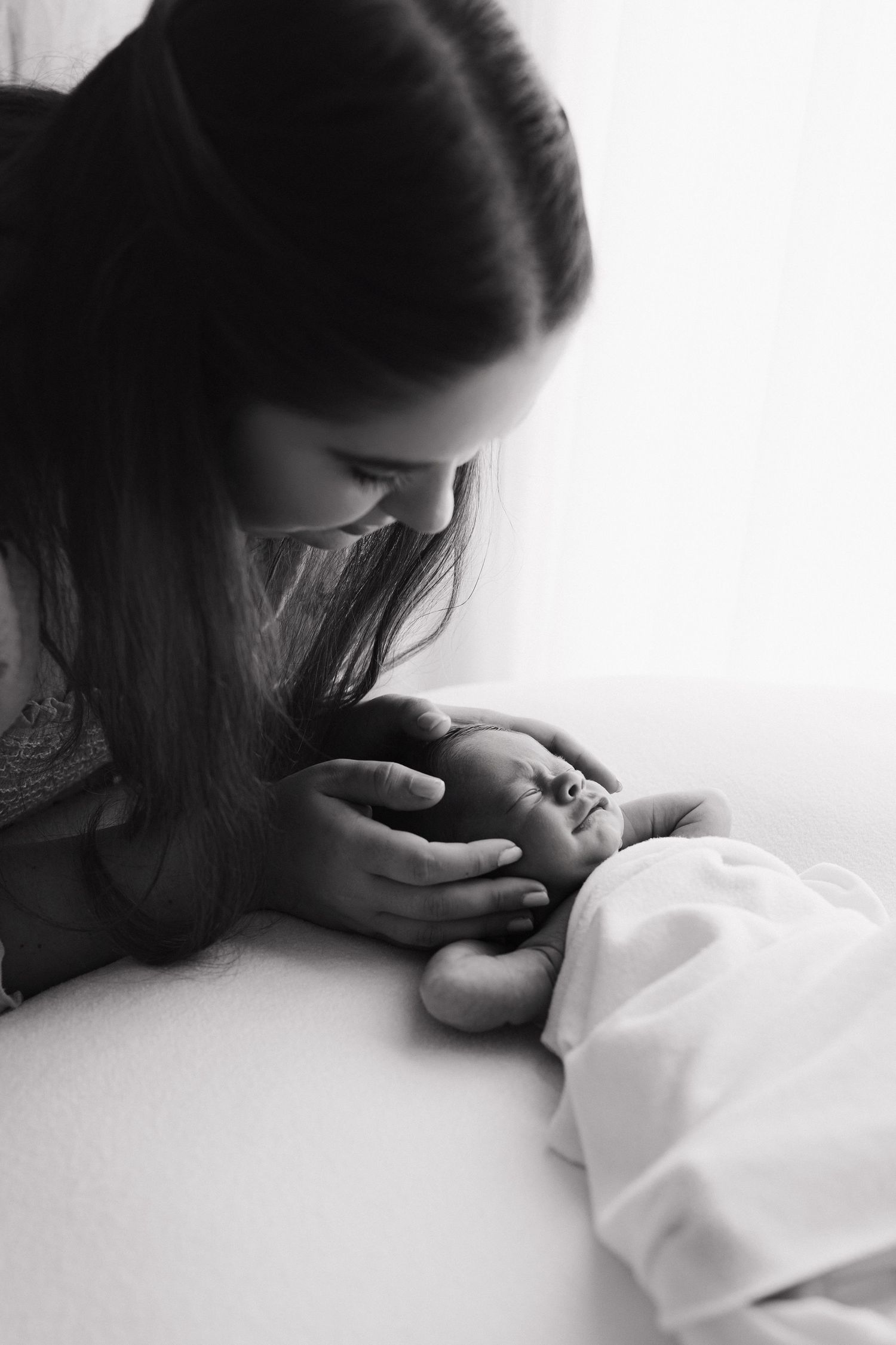 Black and white photo of a young mother tenderly cradling her newborn baby's head while leaning in close.
