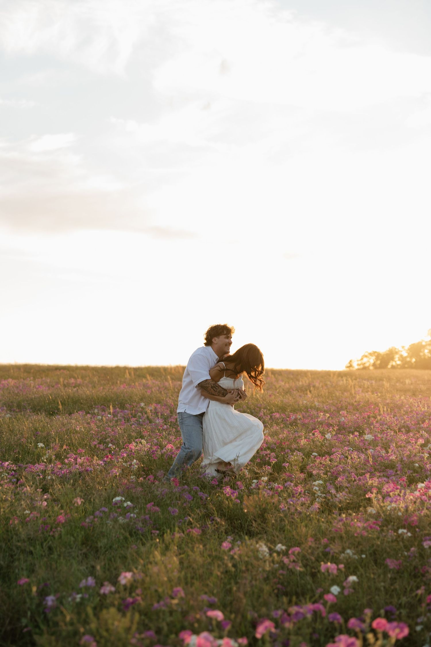 Couple dancing romantically in a blooming wildflower field at golden sunset, wearing white outfits.