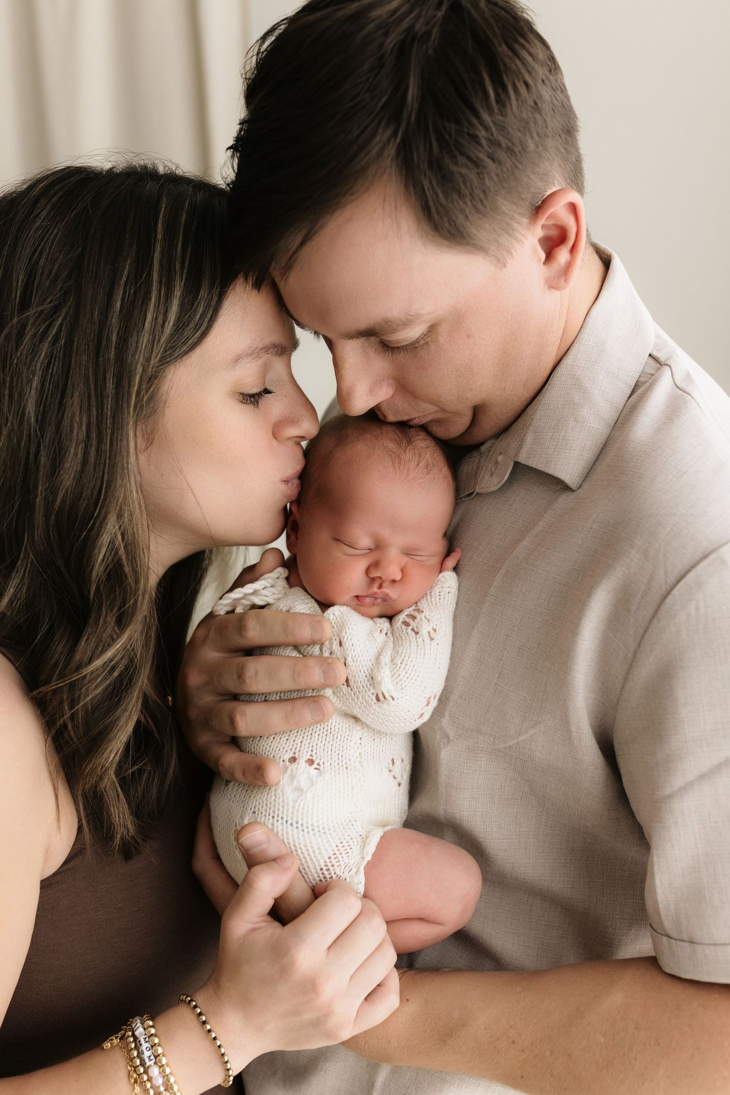 Parents lovingly kiss their sleeping newborn baby wrapped in a white knit swaddle during a studio newborn photo session.