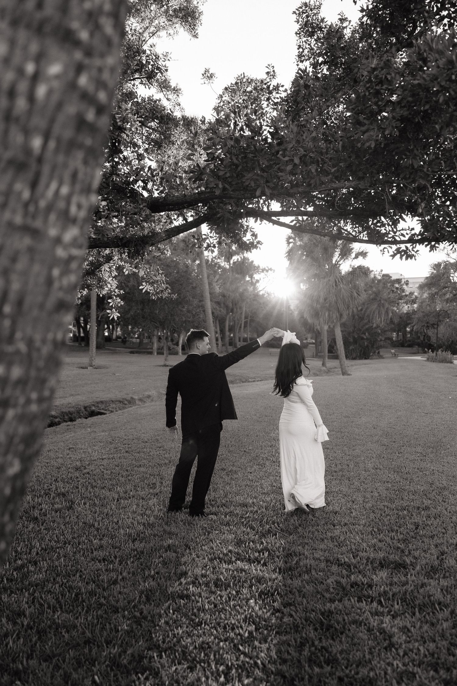 Black and white wedding couple silhouettes dance in a grassy field at sunset with trees in the background.