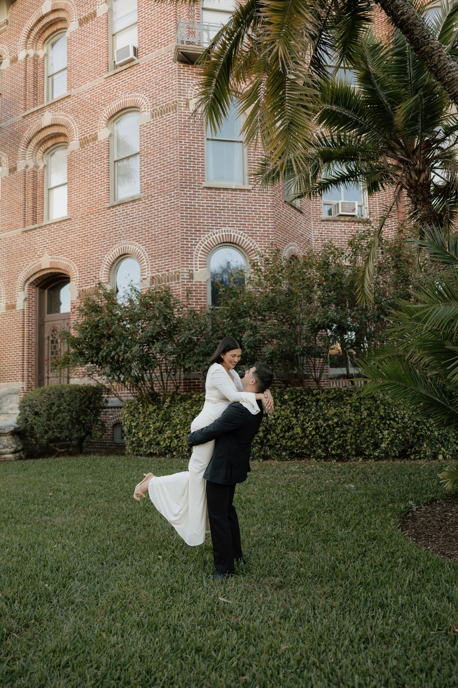 A couple in formal wedding attire dances and spins on a grassy lawn in front of a brick building.