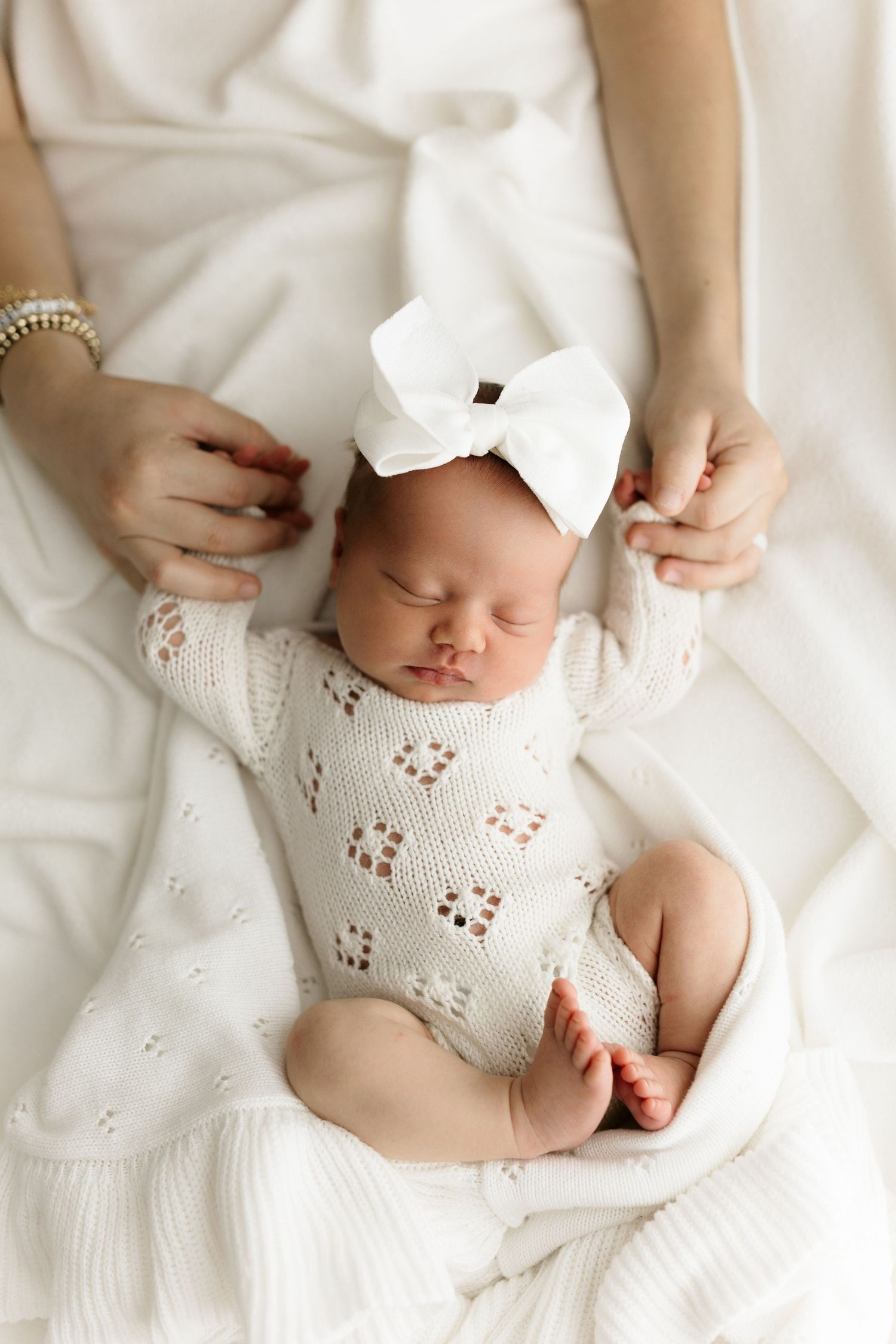 Sleeping newborn baby girl in white knit romper and bow headband cradled in mother's hands on white blanket.