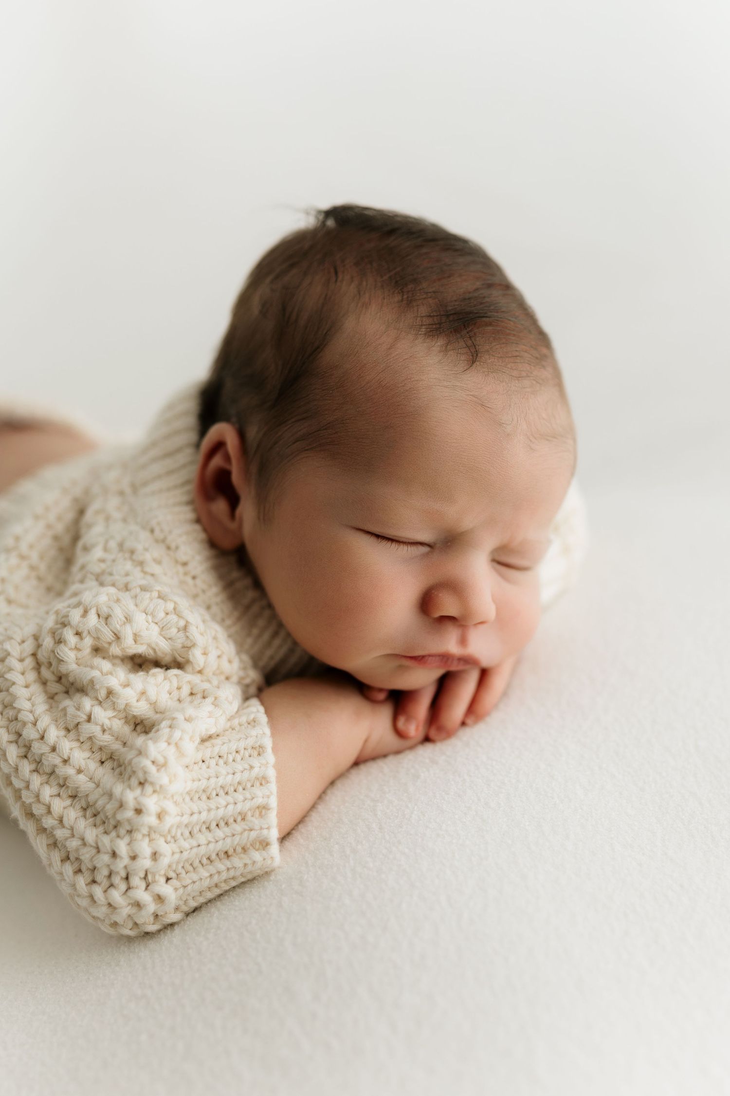 Peaceful newborn baby sleeping in cream knit sweater, resting chin on tiny hands against white background.