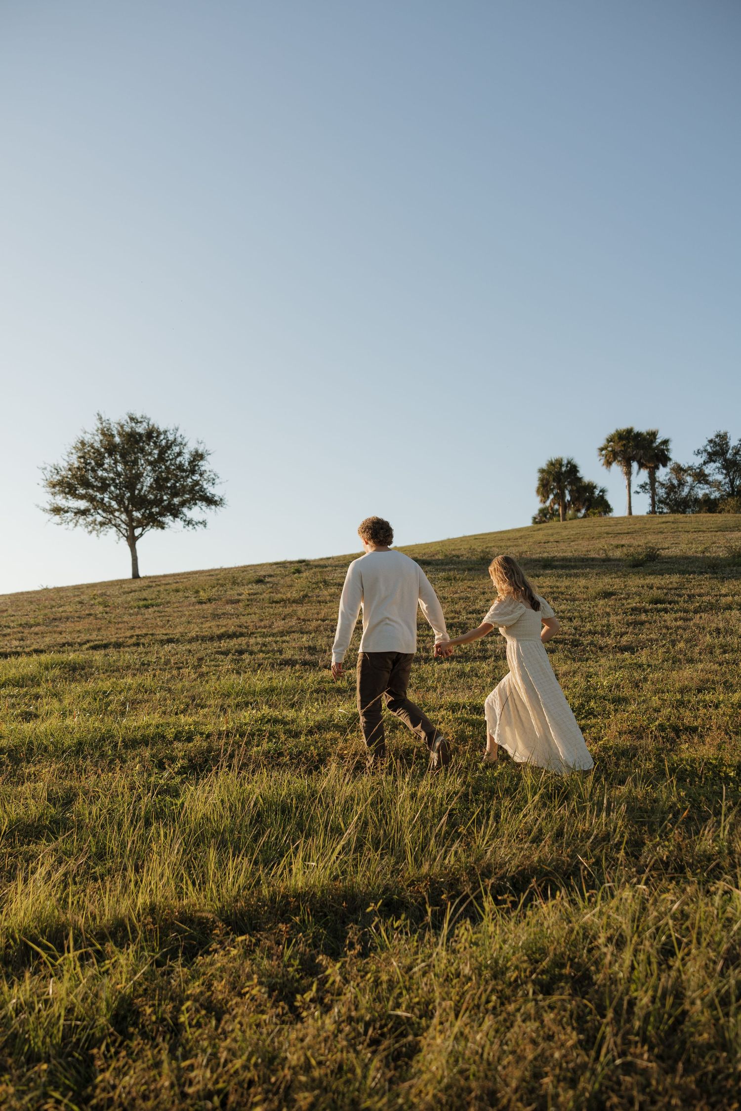 Couple walking up grassy hill at sunset with trees in background.