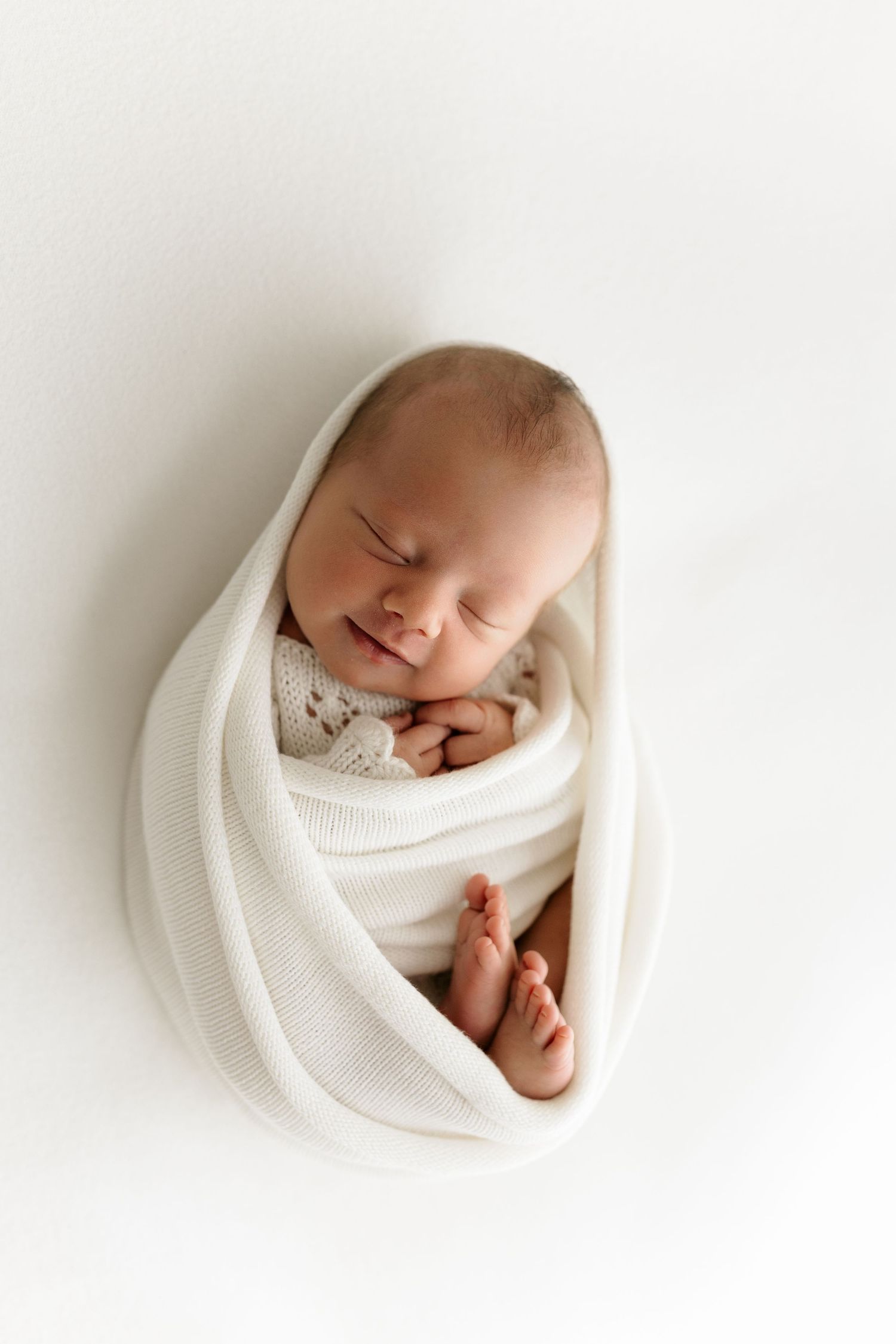 Peaceful newborn baby swaddled in white wrap, sleeping with tiny toes peeking out on white background.