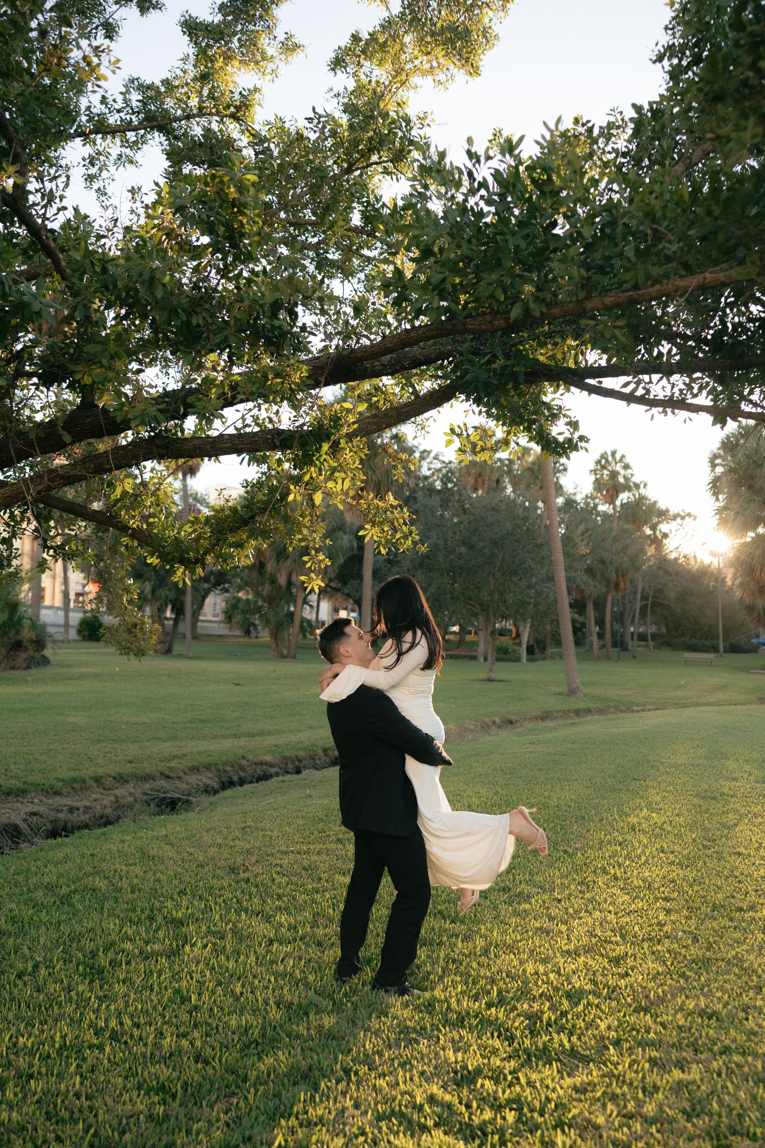 A couple dances and spins together in a grassy field at sunset under a large tree branch.