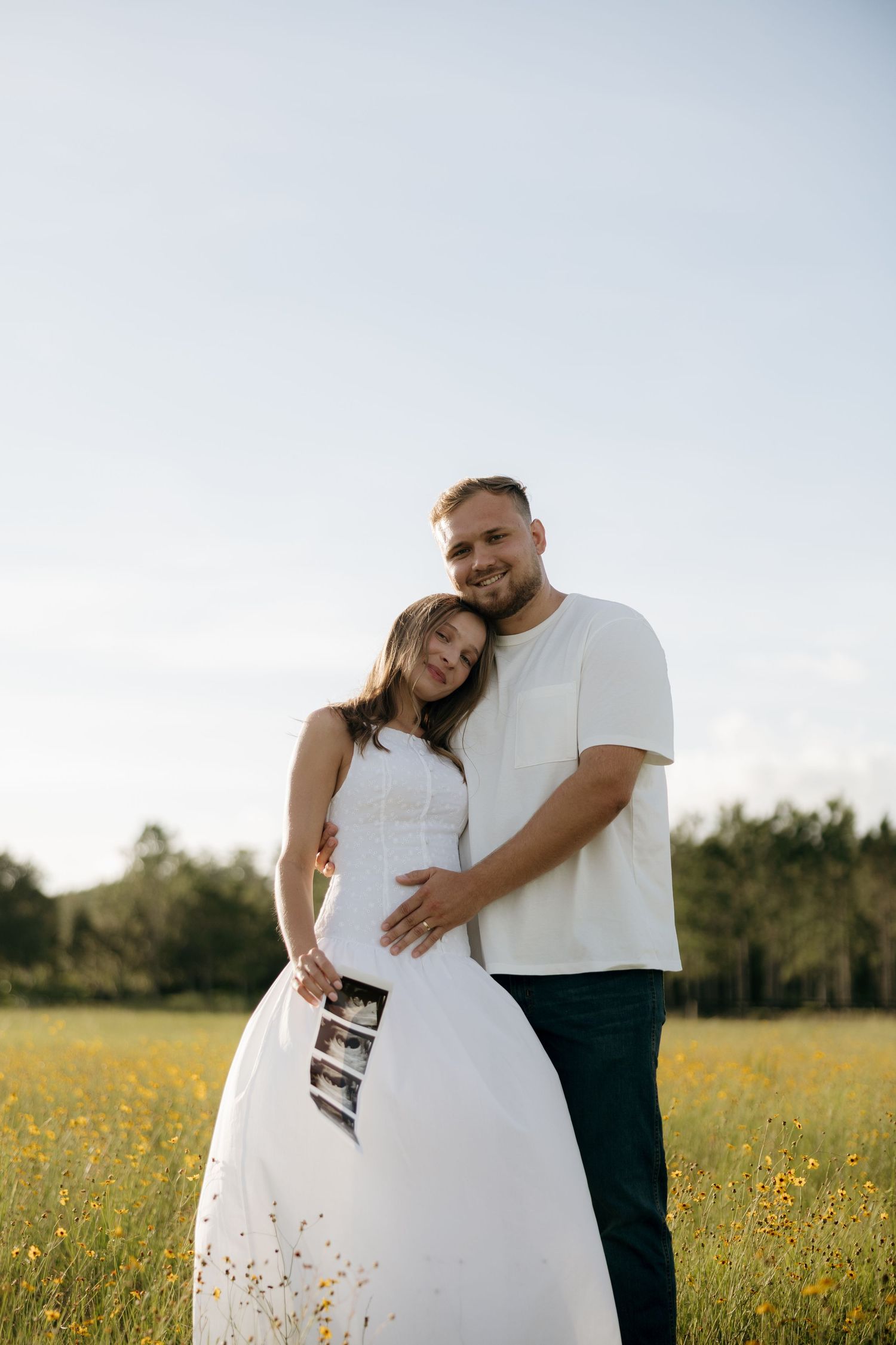 Happy couple embracing in a sunny flower field, woman in white dress holding ultrasound photos during maternity shoot.