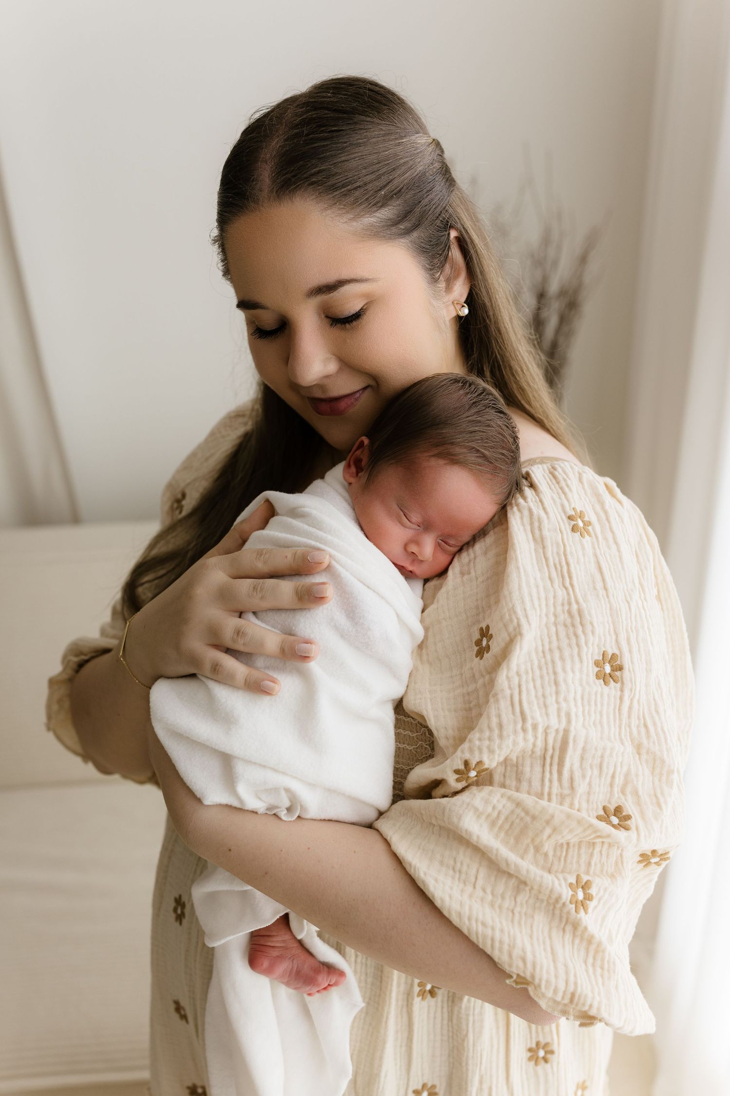 A young mother in a floral dress tenderly holds her sleeping newborn baby wrapped in a white swaddle blanket.
