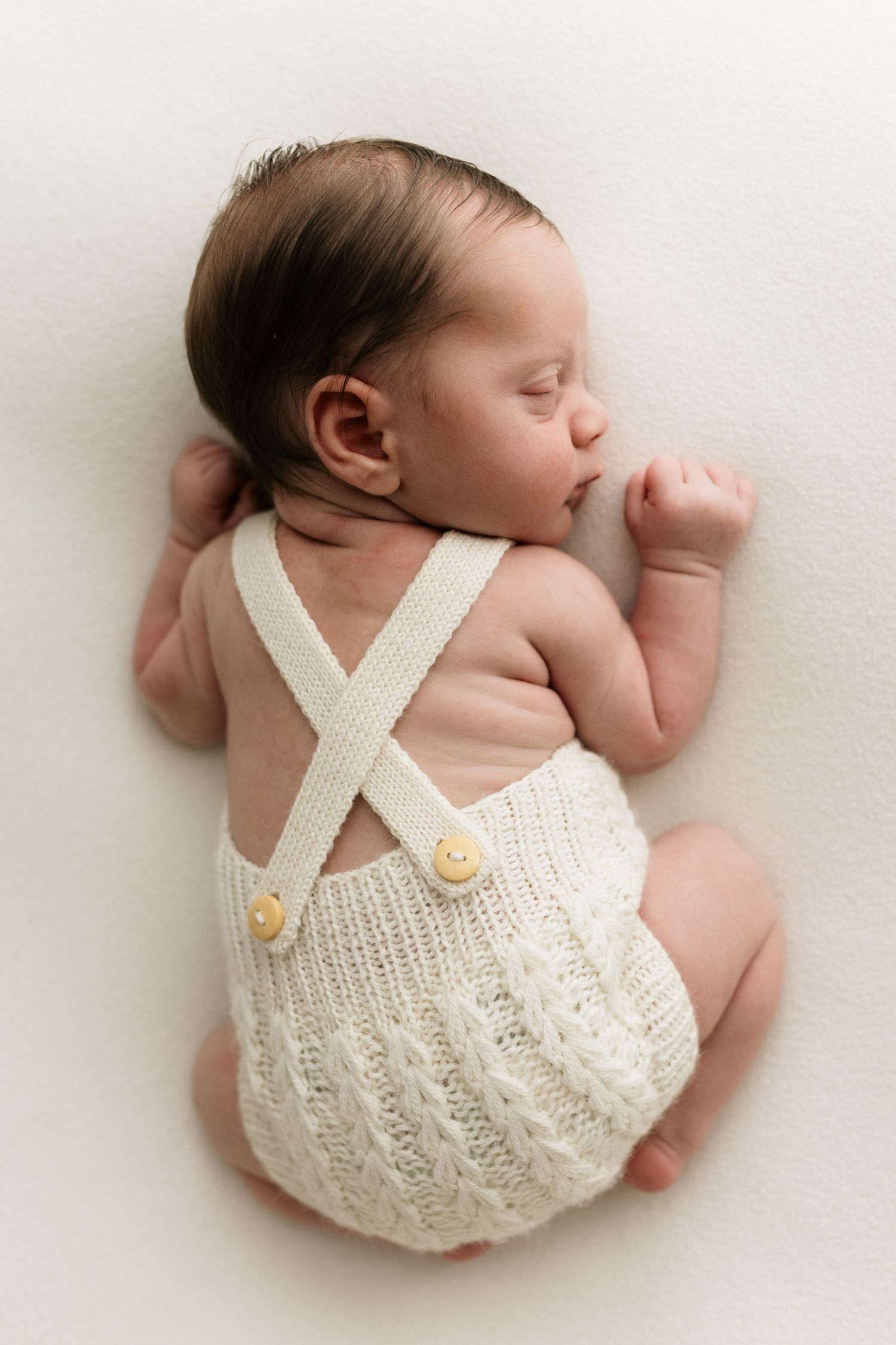 Sleeping newborn baby wearing a cream knit romper with suspenders, lying on a white background.