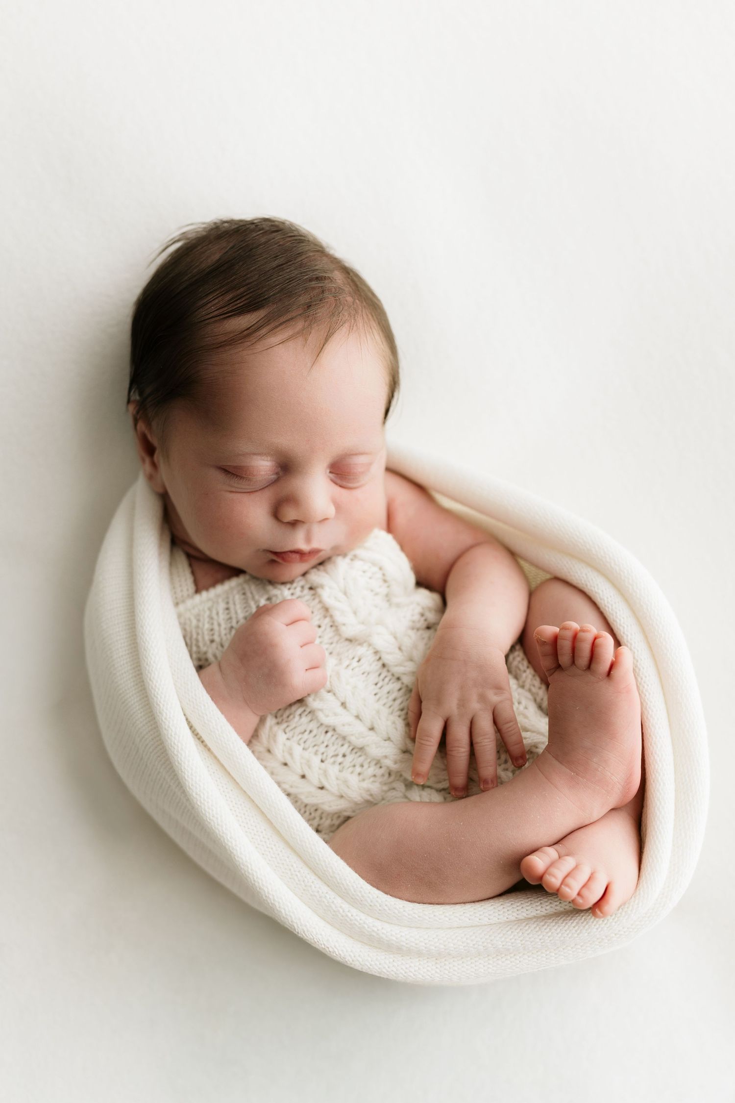 Peaceful newborn baby sleeping curled in a white knit wrap on a soft white background.