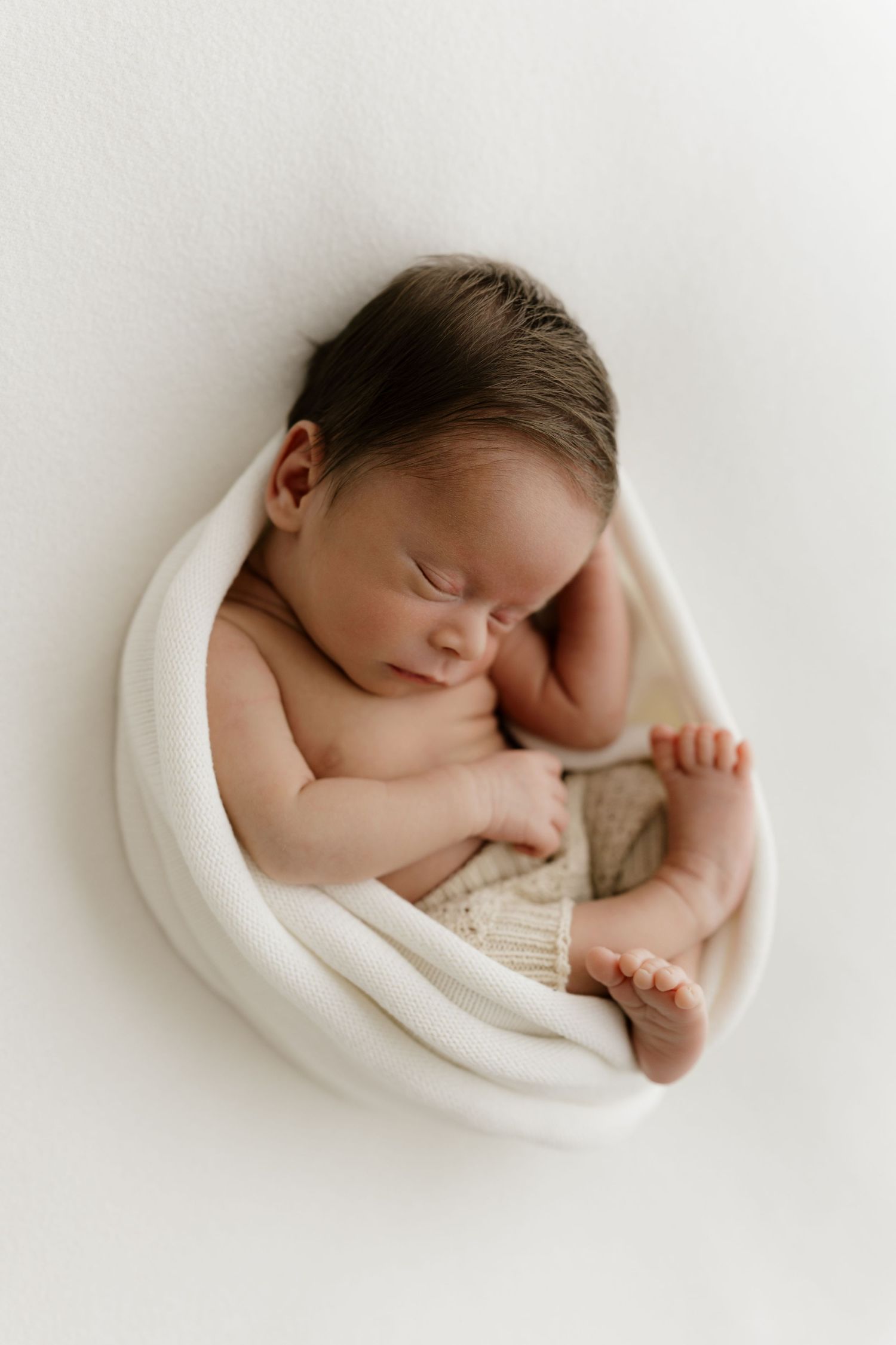 Sleeping newborn baby wrapped in white swaddle wearing knit shorts, curled in fetal position on white background.