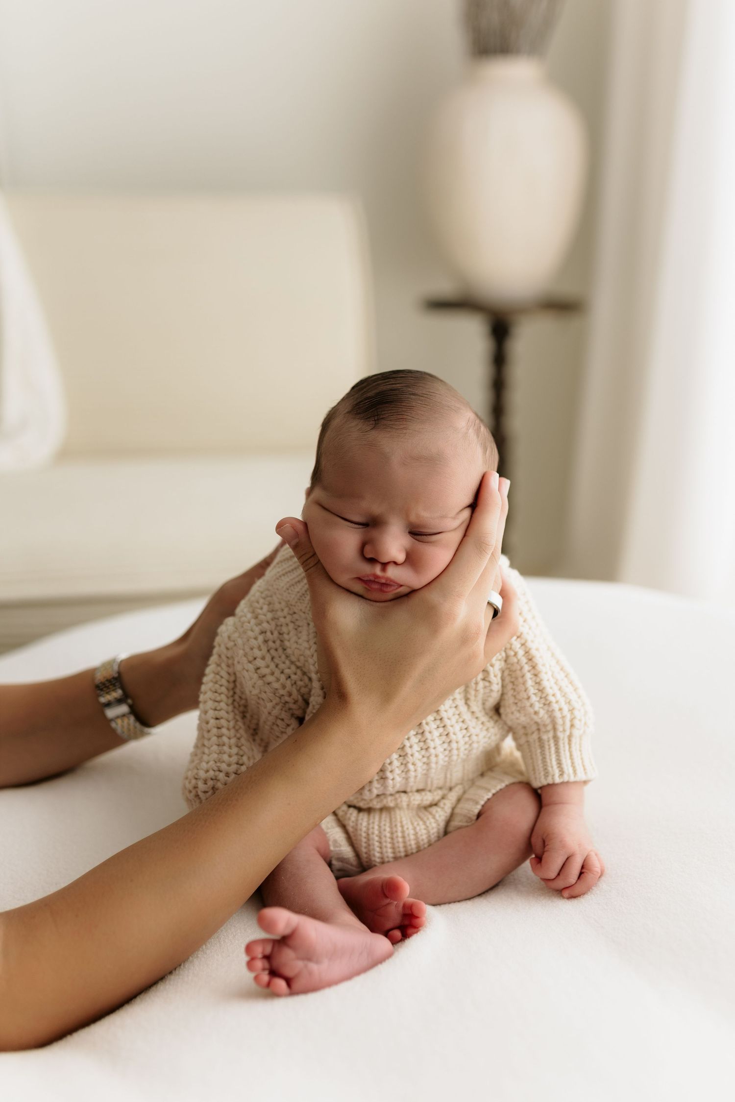 A mother gently cradles her sleeping newborn baby dressed in a cream knit romper on a white surface.
