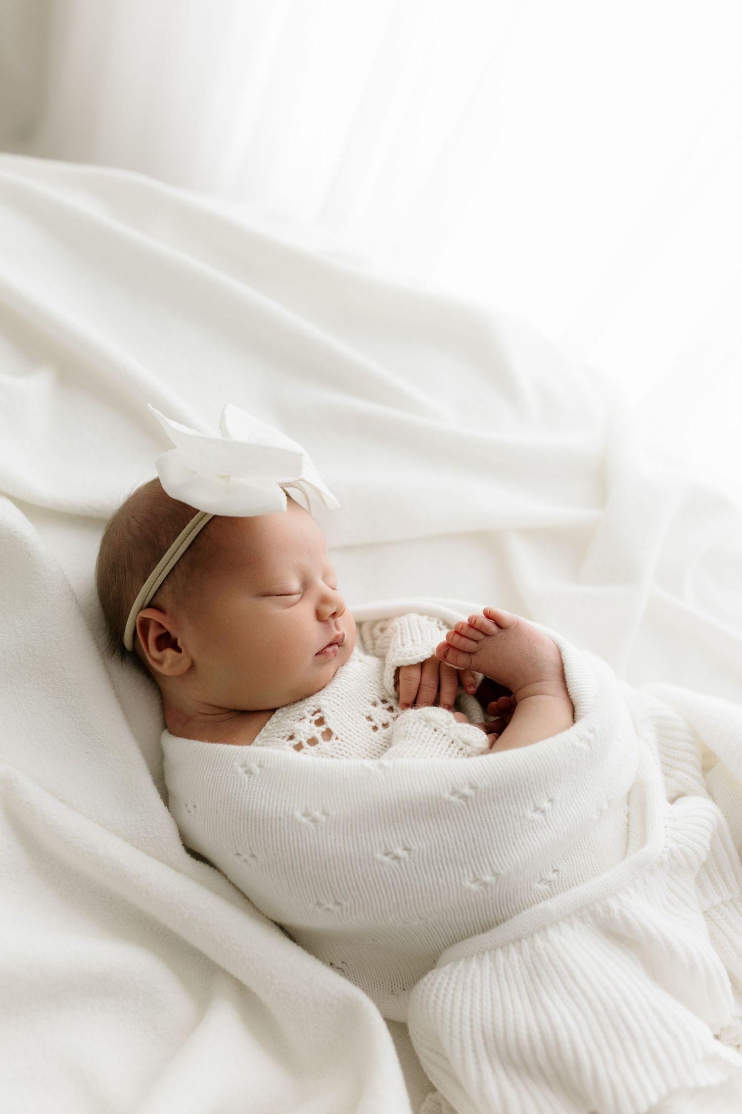 Peaceful newborn baby girl sleeping wrapped in white knit blanket with white bow headband on soft white bedding.