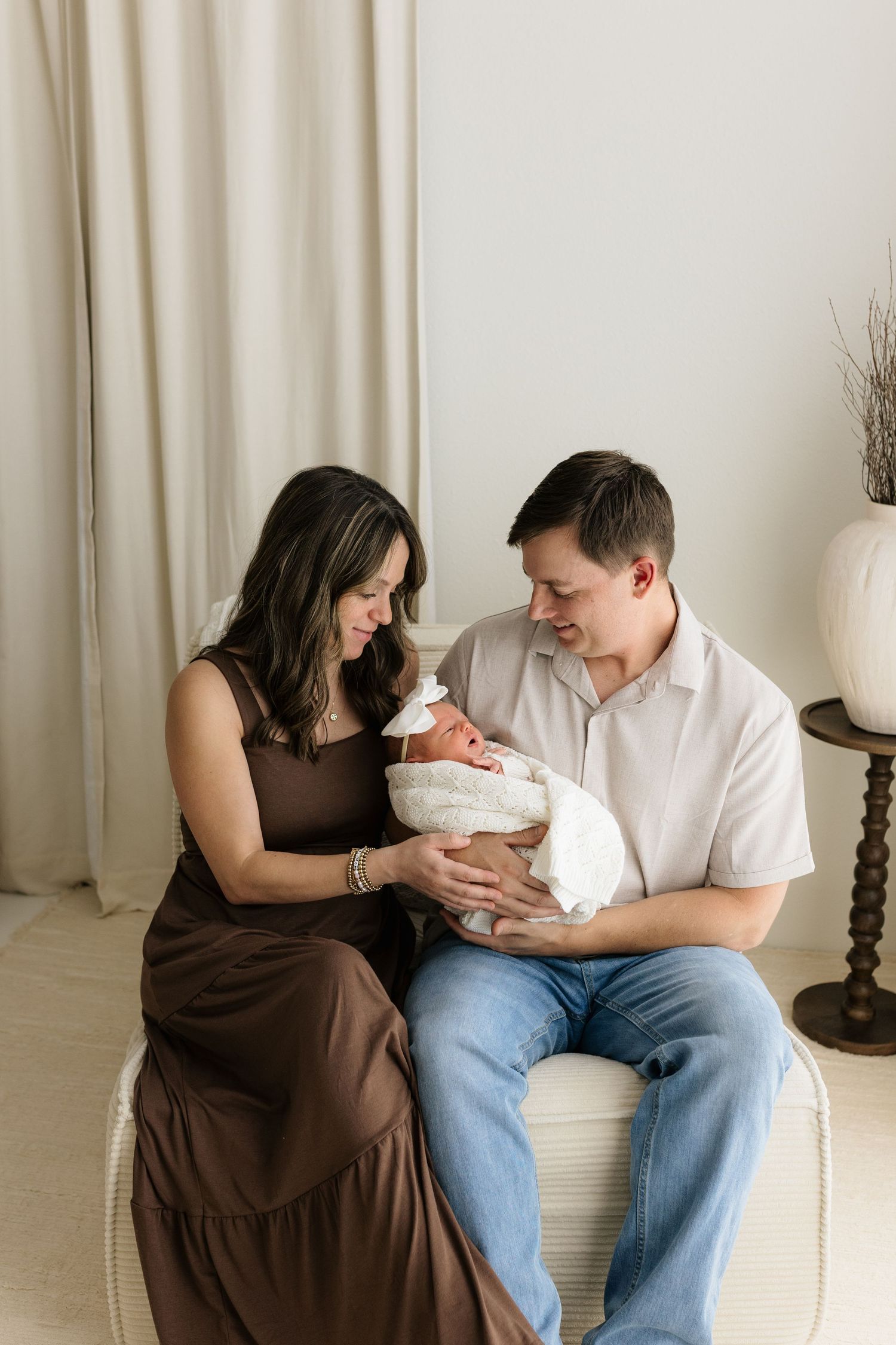 A couple in neutral tones sits together holding their swaddled newborn baby in a bright, minimalist studio setting.