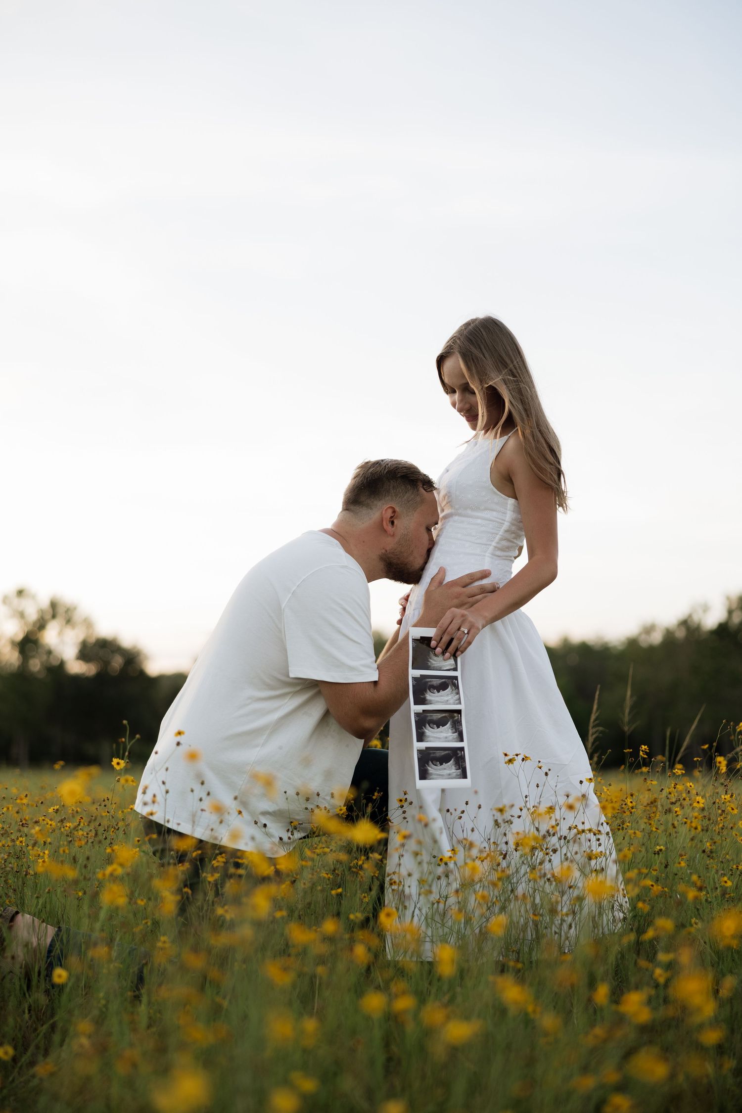 Man kissing pregnant woman's belly in a sunny yellow wildflower field, holding ultrasound photos.