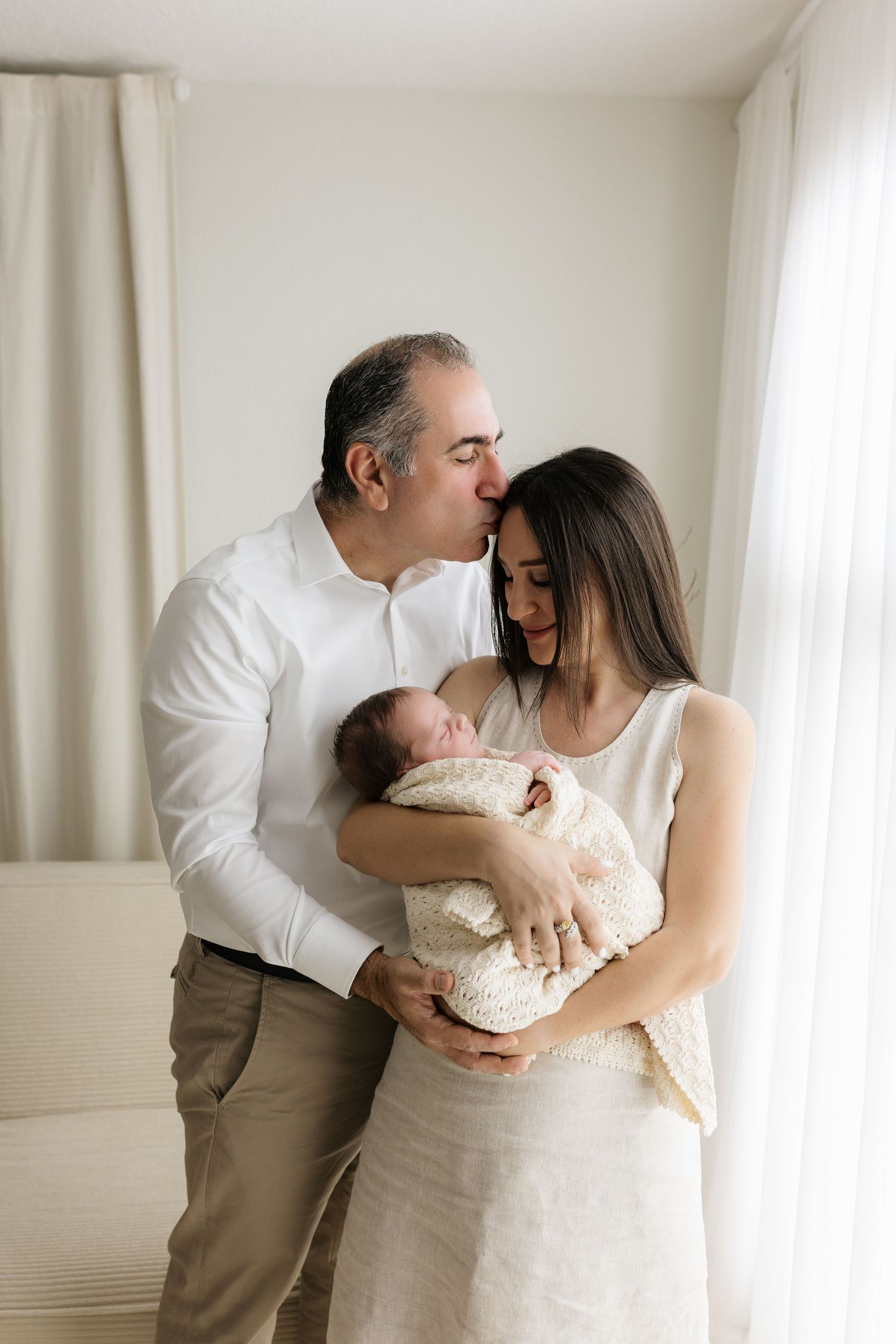 A couple in white clothing tenderly holds their newborn baby, with the man kissing the woman's head indoors.