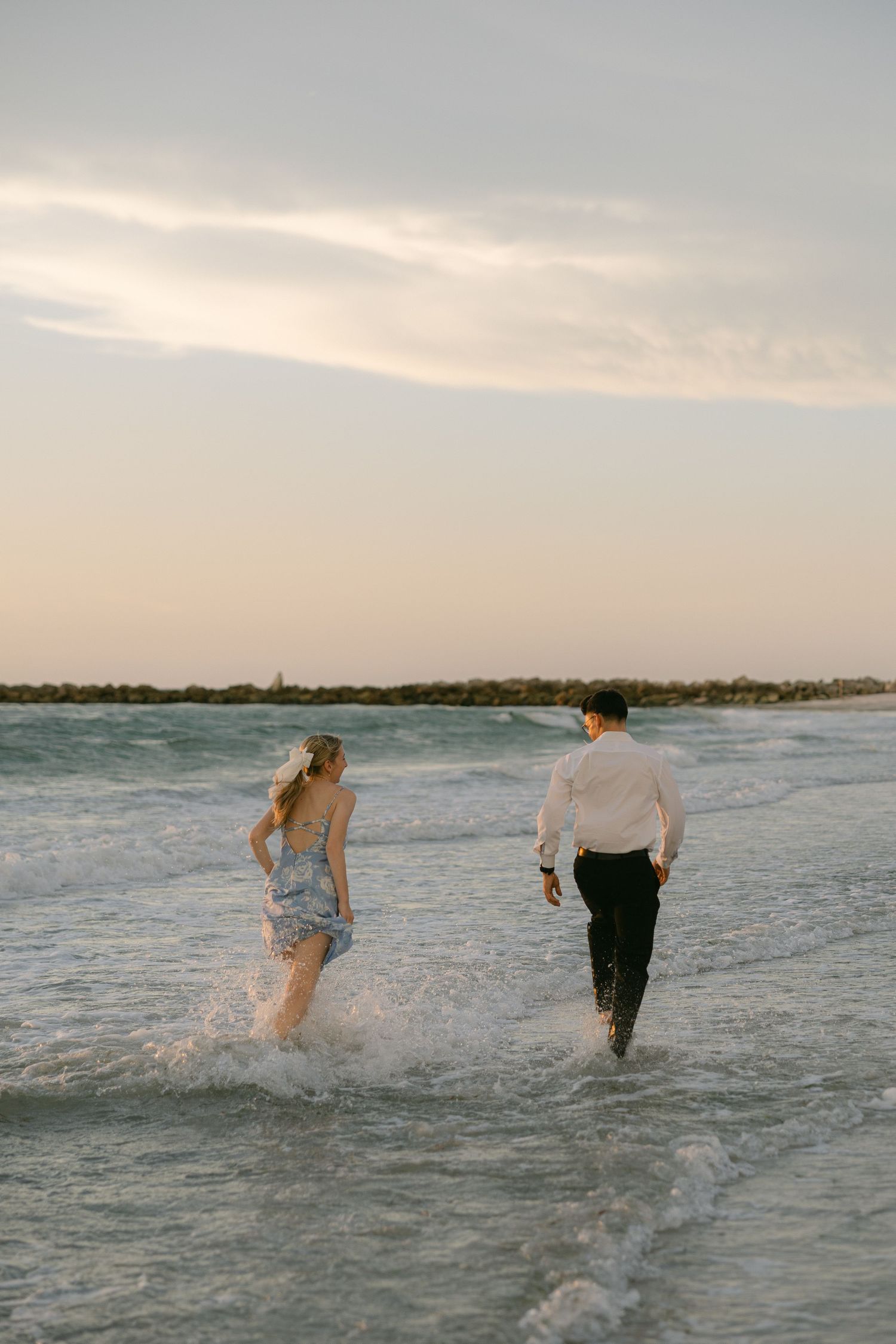Two people splash and run through shallow ocean waves at sunset along a peaceful beach shoreline.
