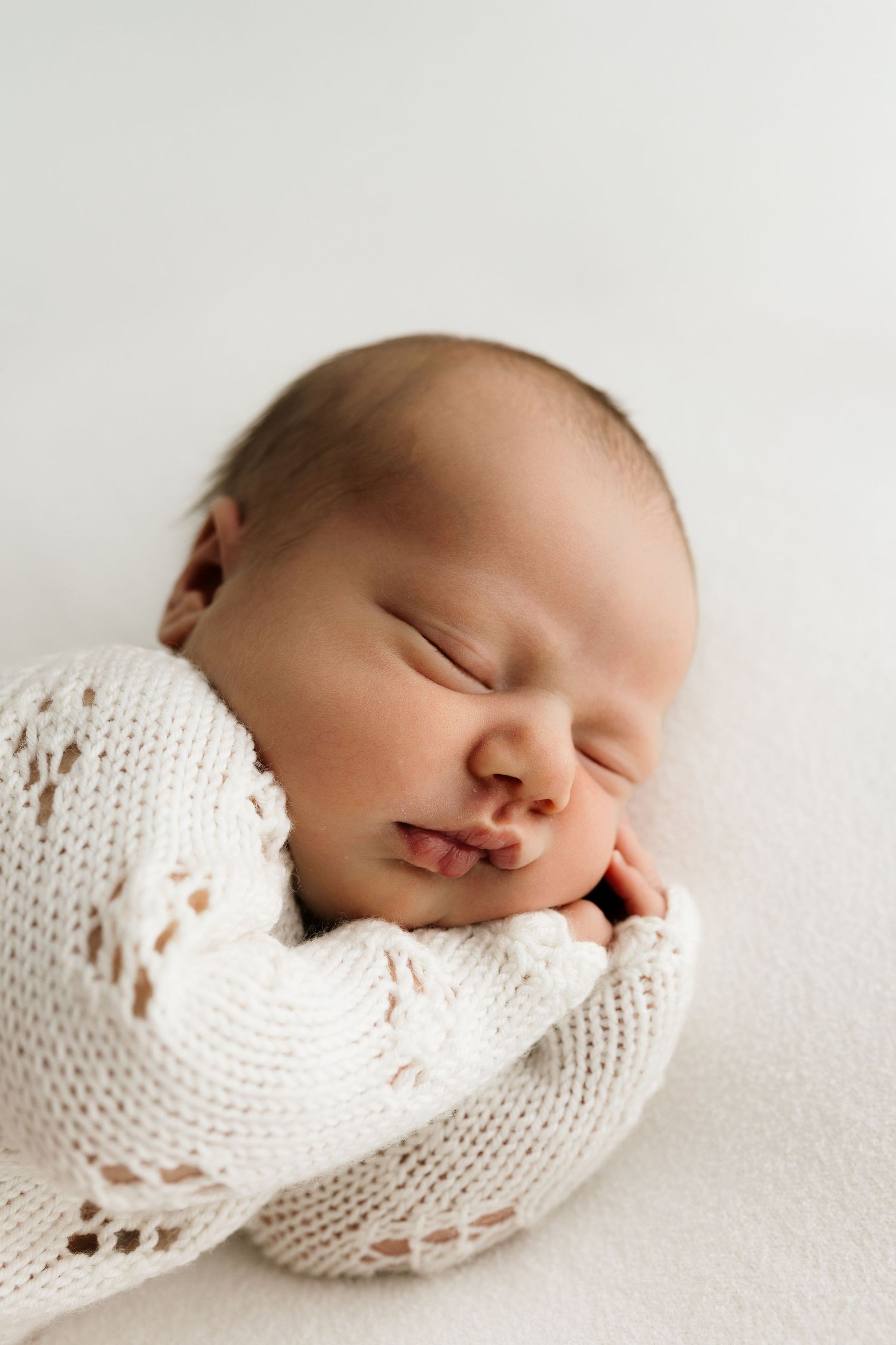 Peaceful newborn baby sleeping curled up in a white knit outfit on a soft white background.