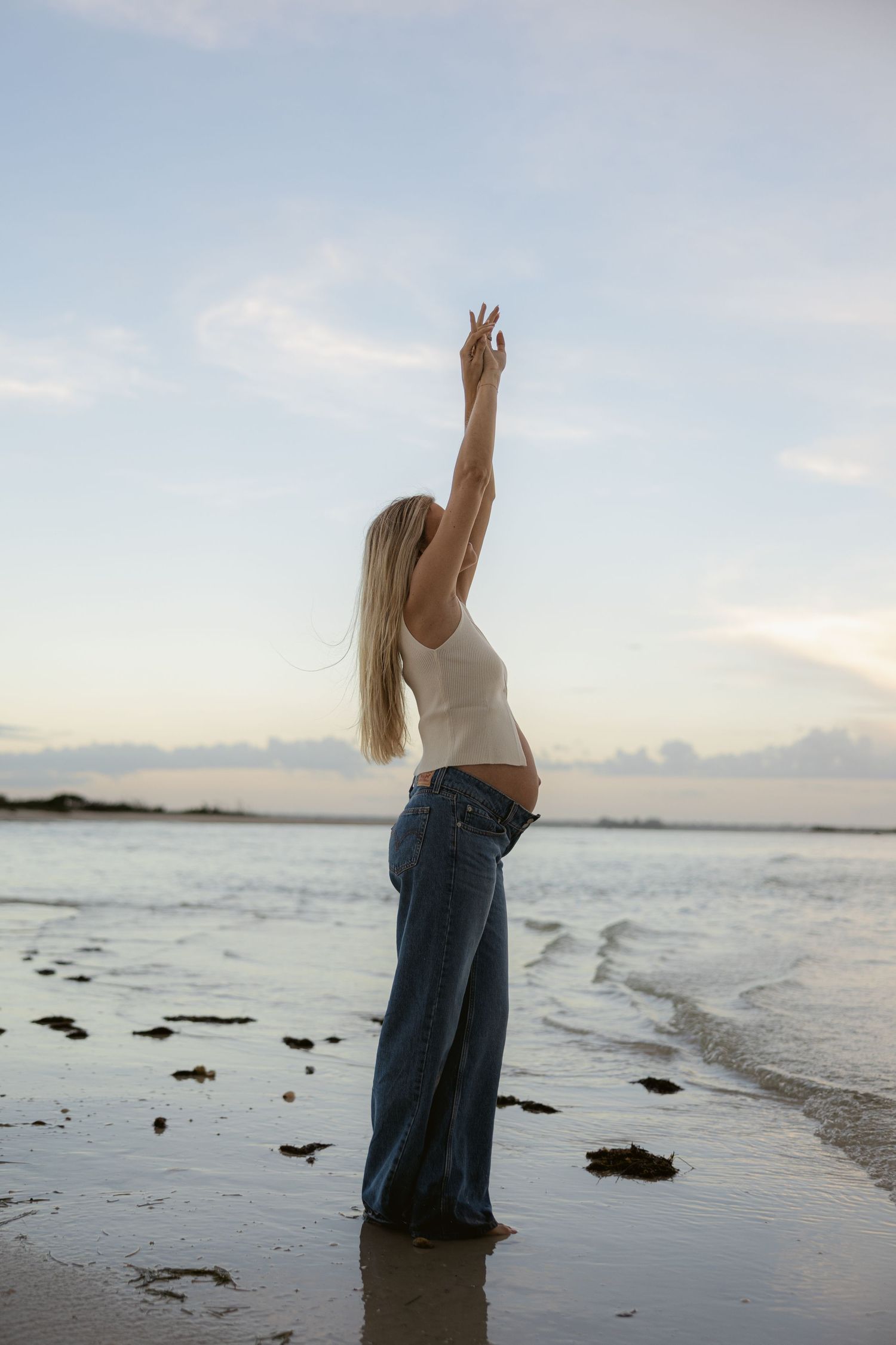 A woman in jeans poses artistically on a beach at sunset with waves in the background.