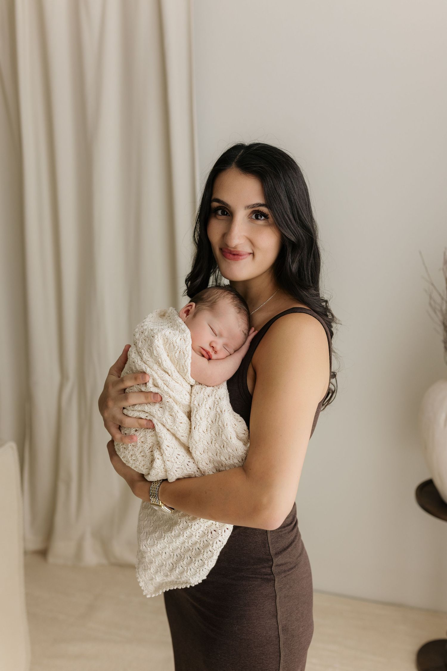 A dark-haired woman in brown holds a sleeping newborn wrapped in a white knit blanket, smiling warmly in a bright studio.