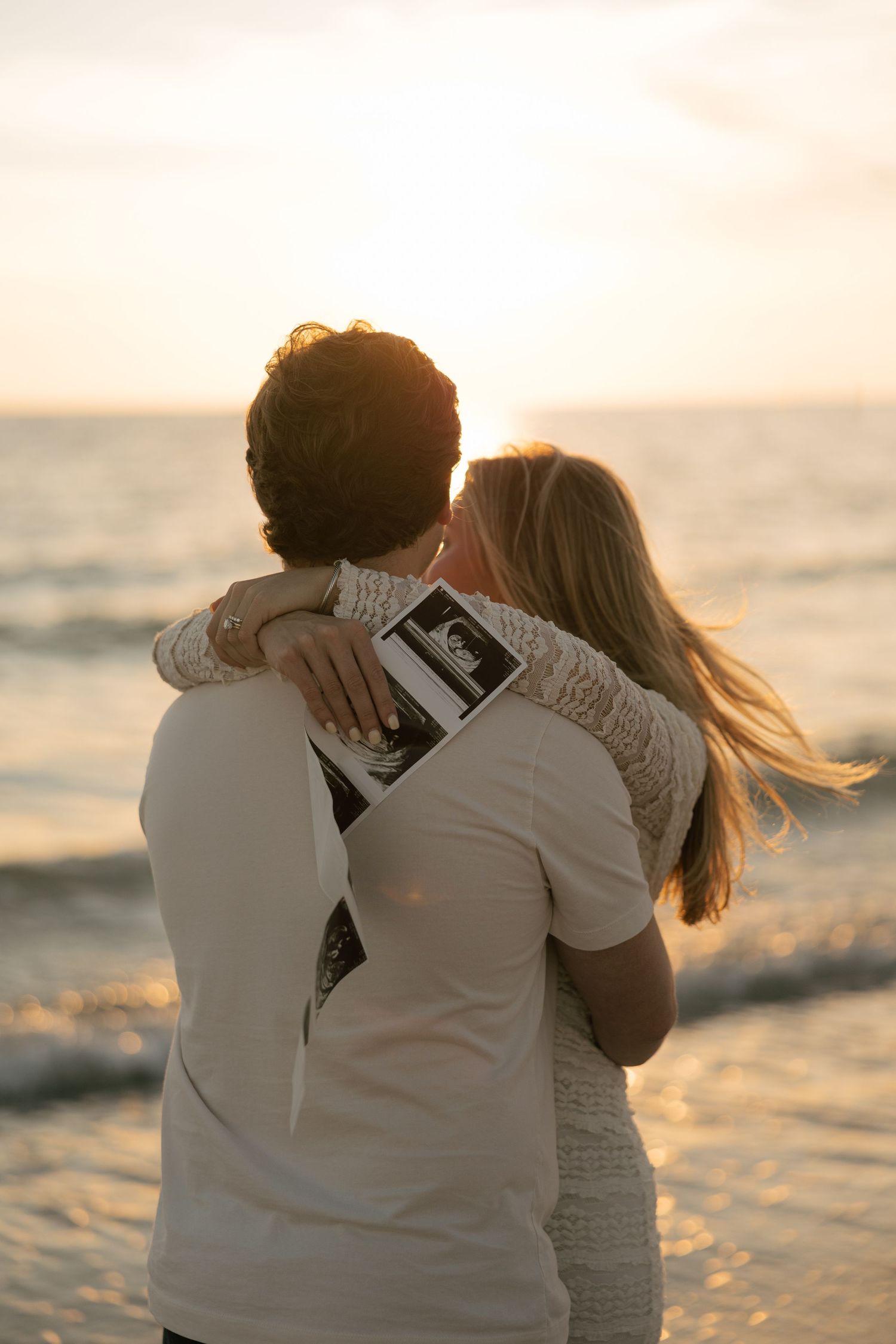 Couple embracing on beach at sunset, holding ultrasound photo, announcing pregnancy with ocean waves in background.