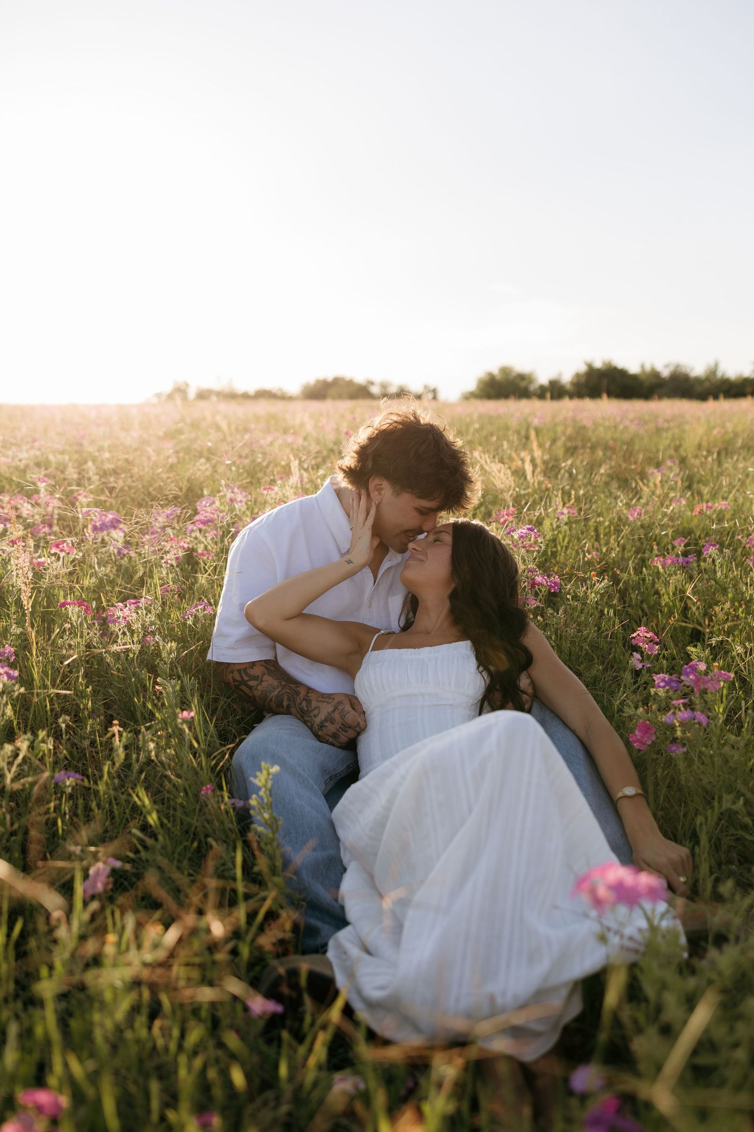 Couple sharing a romantic almost-kiss in a sunny wildflower field, woman in white dress, man in jeans and white shirt.