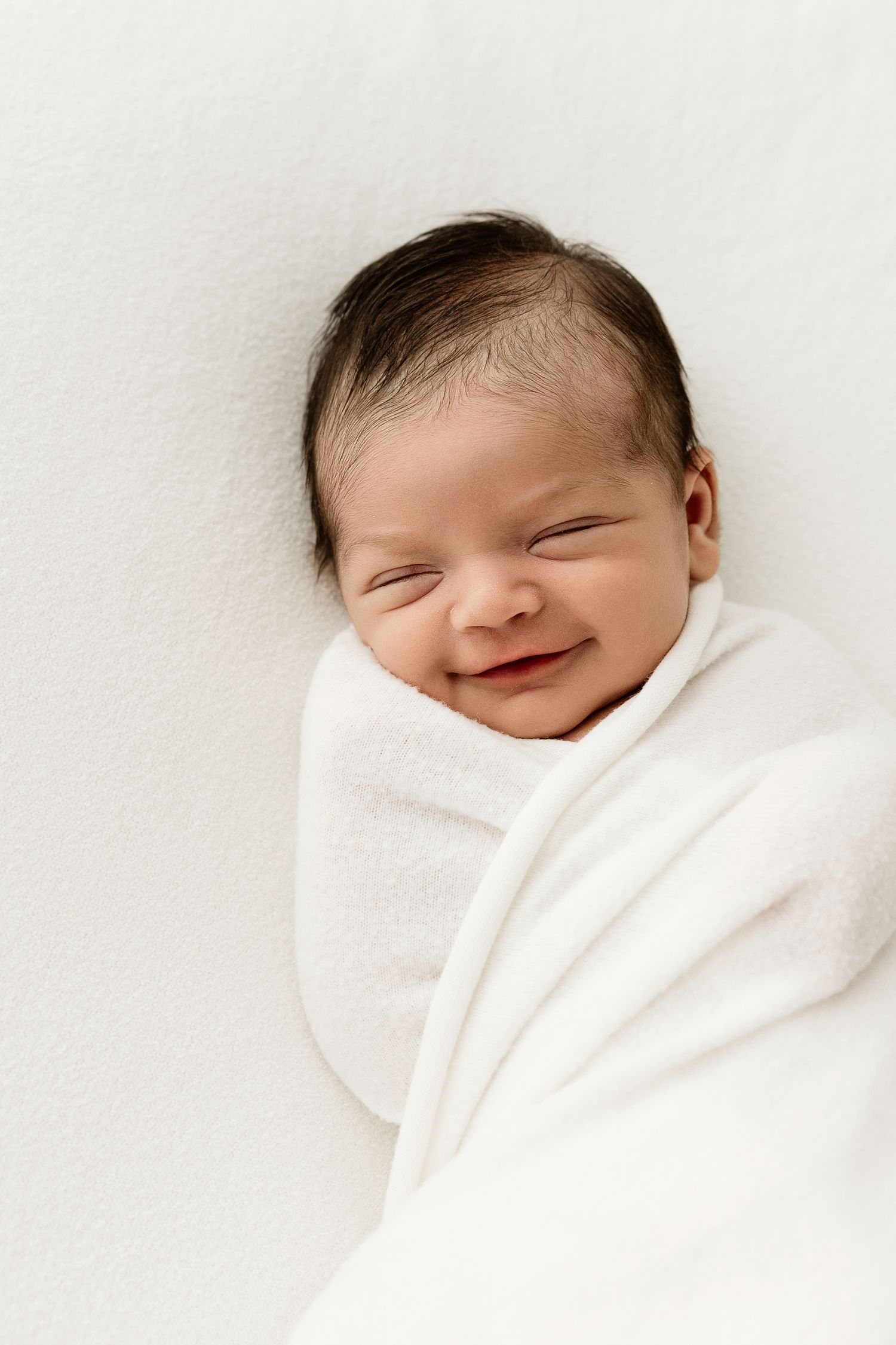 Smiling newborn baby swaddled in a white blanket lying on a soft white background.
